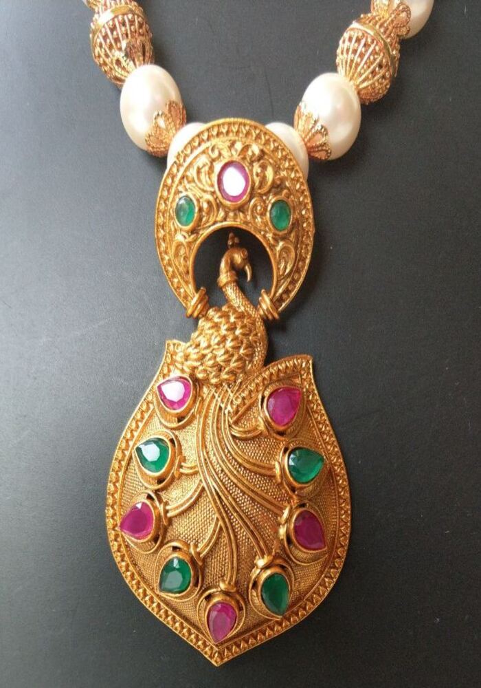 Detailed, colorful peacock-inspired pendant with intricate golden filigree and vibrant gemstones, set against a dark background, showcasing the ornate temple jewelry design from the Mugdha Jewellery Store.