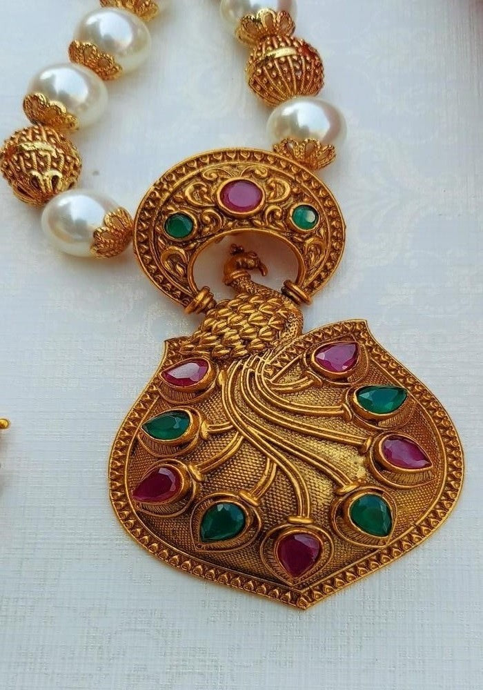 Ornate gold-toned peacock pendant with colorful gemstones and pearls on a necklace, displayed in a Mugdha Jewellery Store product image.