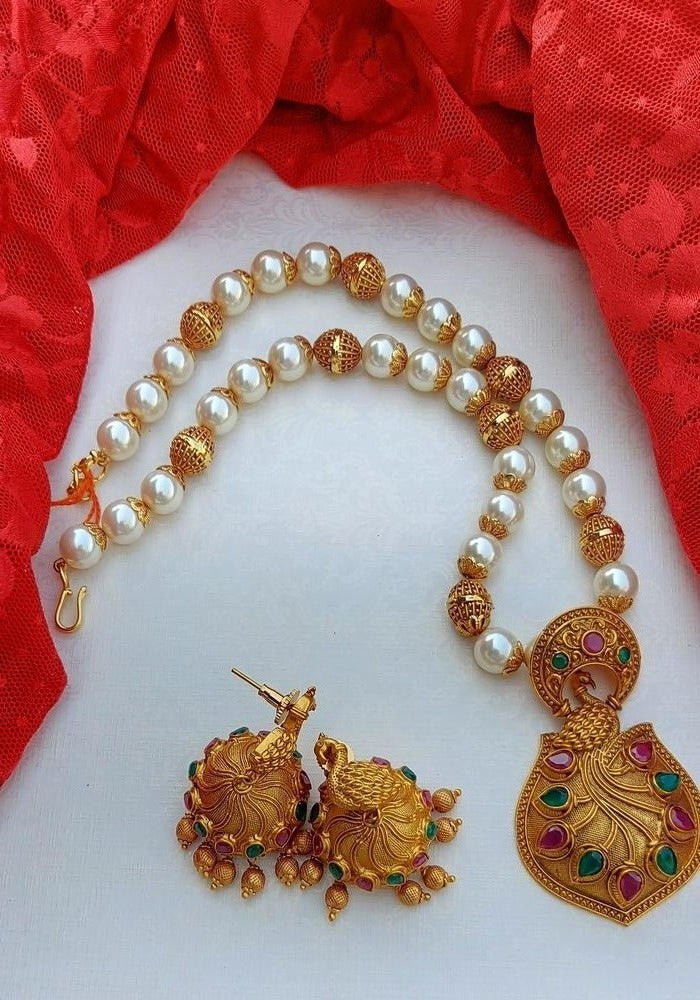Elegant pearl and gold-toned temple jewelry set featuring a dancing peacock pendant on a pearl necklace, surrounded by a vibrant red fabric backdrop.