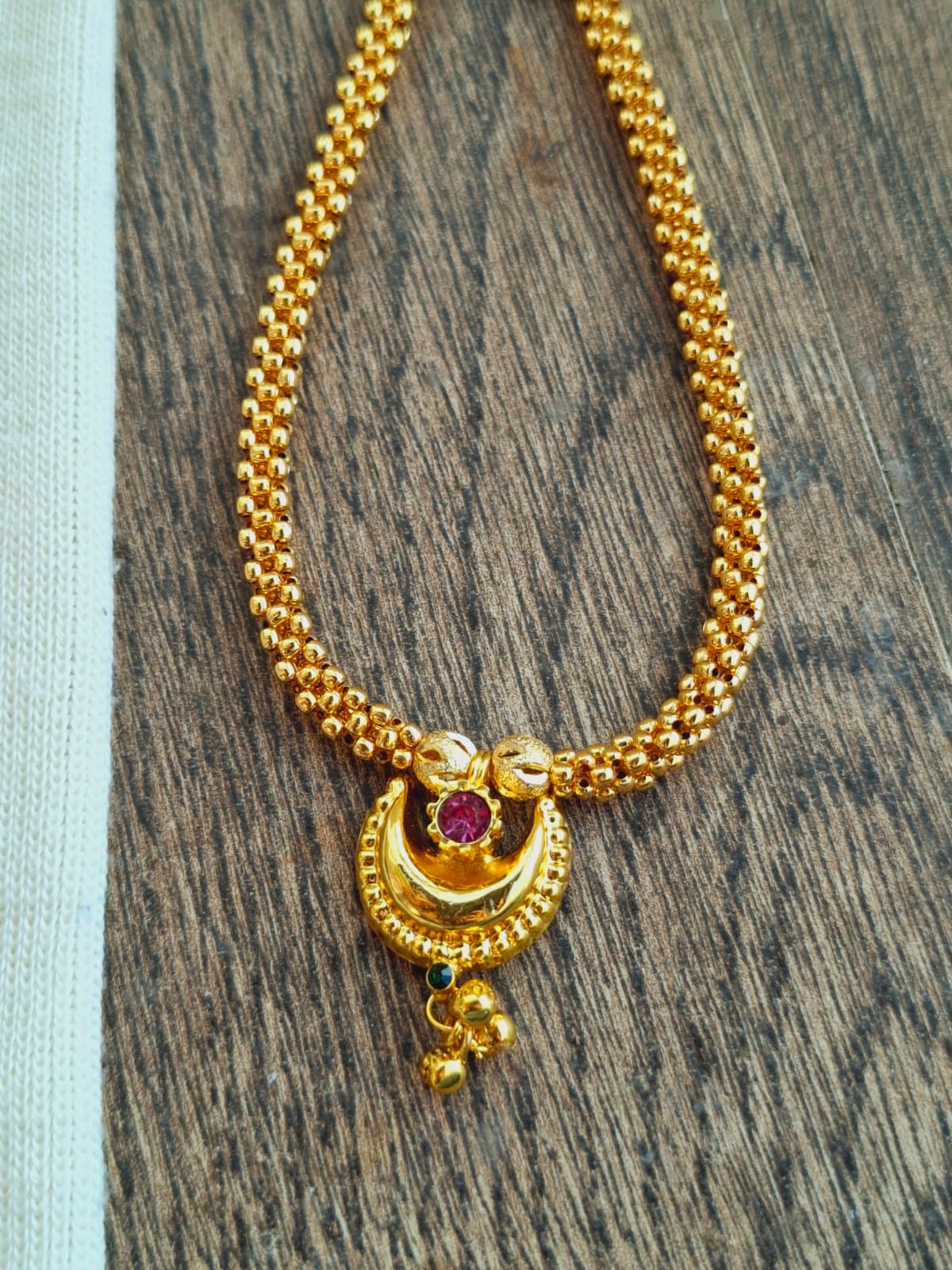 Elegant gold beaded necklace with a heart-shaped pendant featuring a ruby gemstone, displayed on a wooden background.
