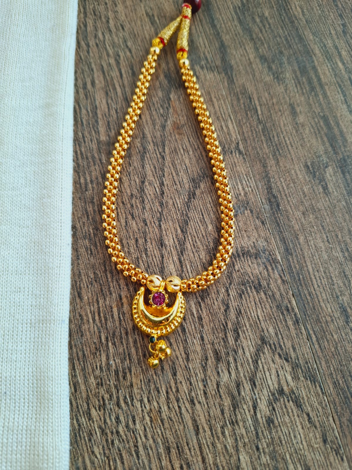 Elegant gold necklace with intricate pendant featuring a crescent moon shape and a red gemstone, placed on a rustic wooden surface.
