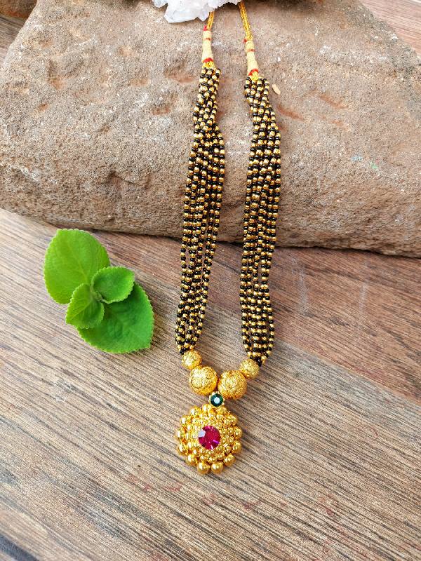 Traditional gold-tone mangalsutra with beaded chain and pendant adorned with red gemstone, placed on a wooden surface with a green leaf.