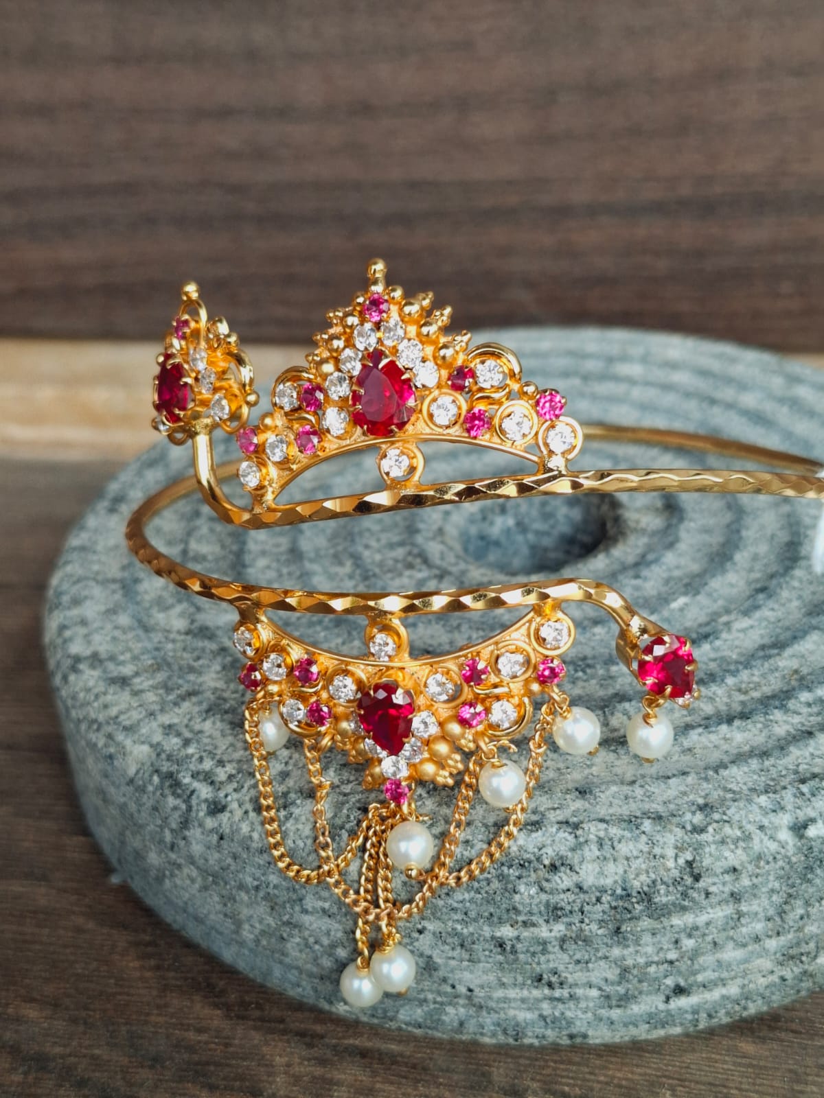Elegant gold-toned bajuband adorned with vibrant red and pink gemstones, placed on a textured gray surface.