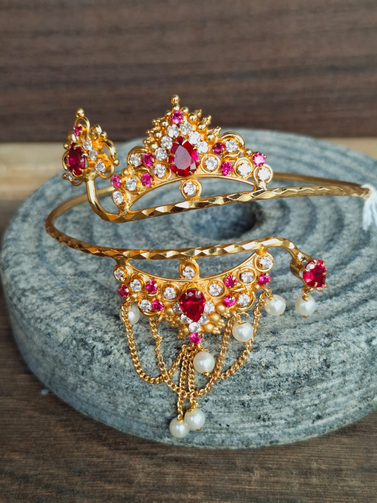 Ornate gold-toned bajuband adorned with vibrant red and pink rhinestones, displayed on a grey textured surface.