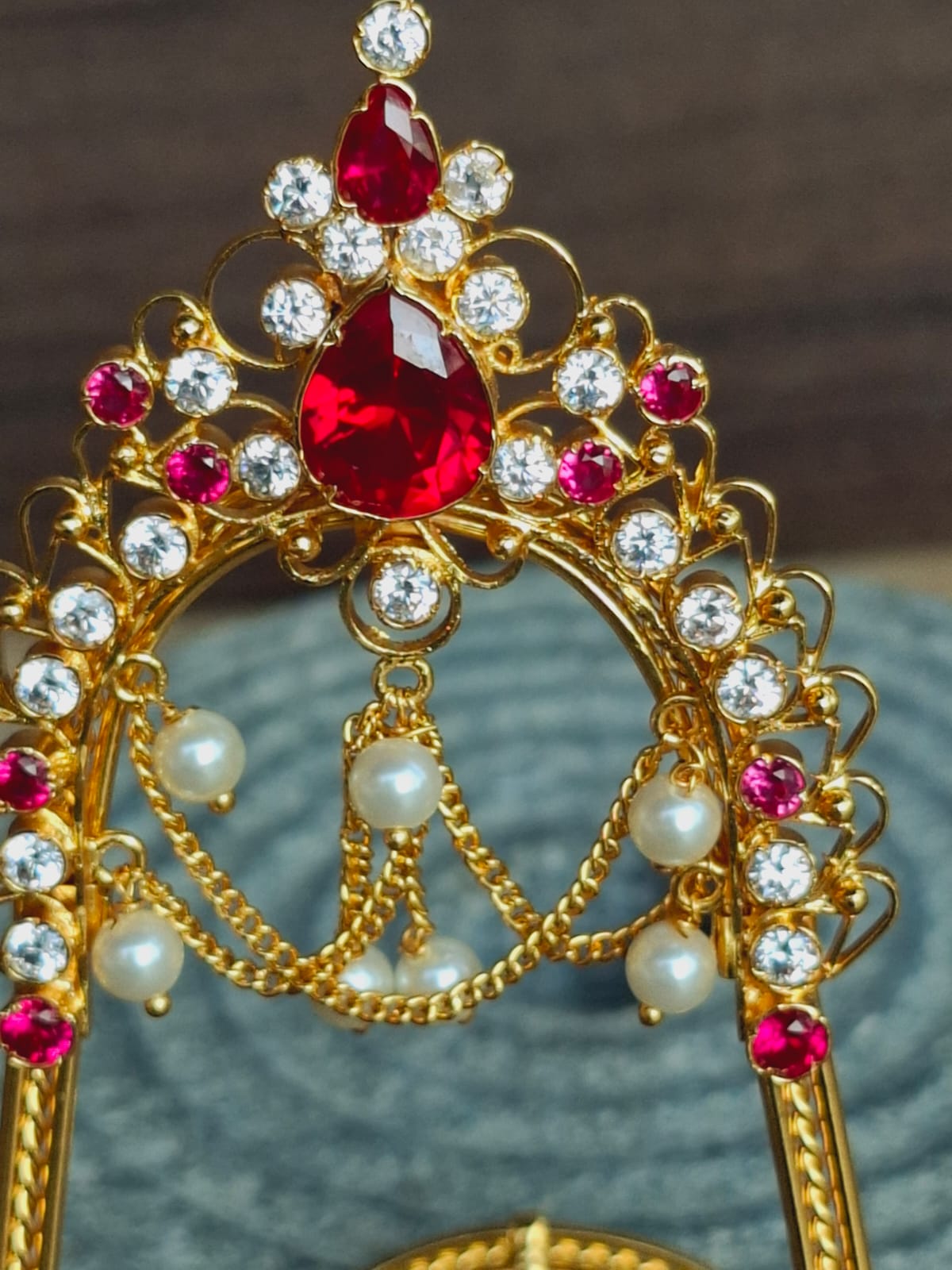 Ornate gold-toned jewelry piece with red and clear gemstones, hanging jewel chains, and pearl accents displayed on a dark background.