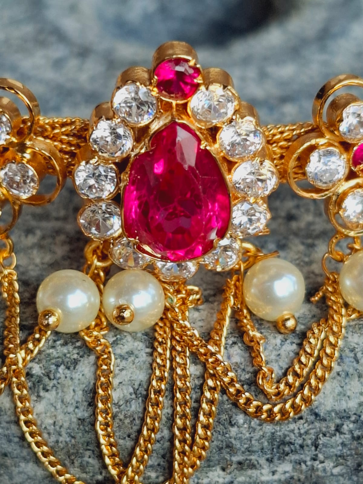 Ornate gold-toned necklace with vibrant red gemstone centerpiece, surrounded by sparkling diamonds and dangling pearl accents, displayed on a textured gray background.