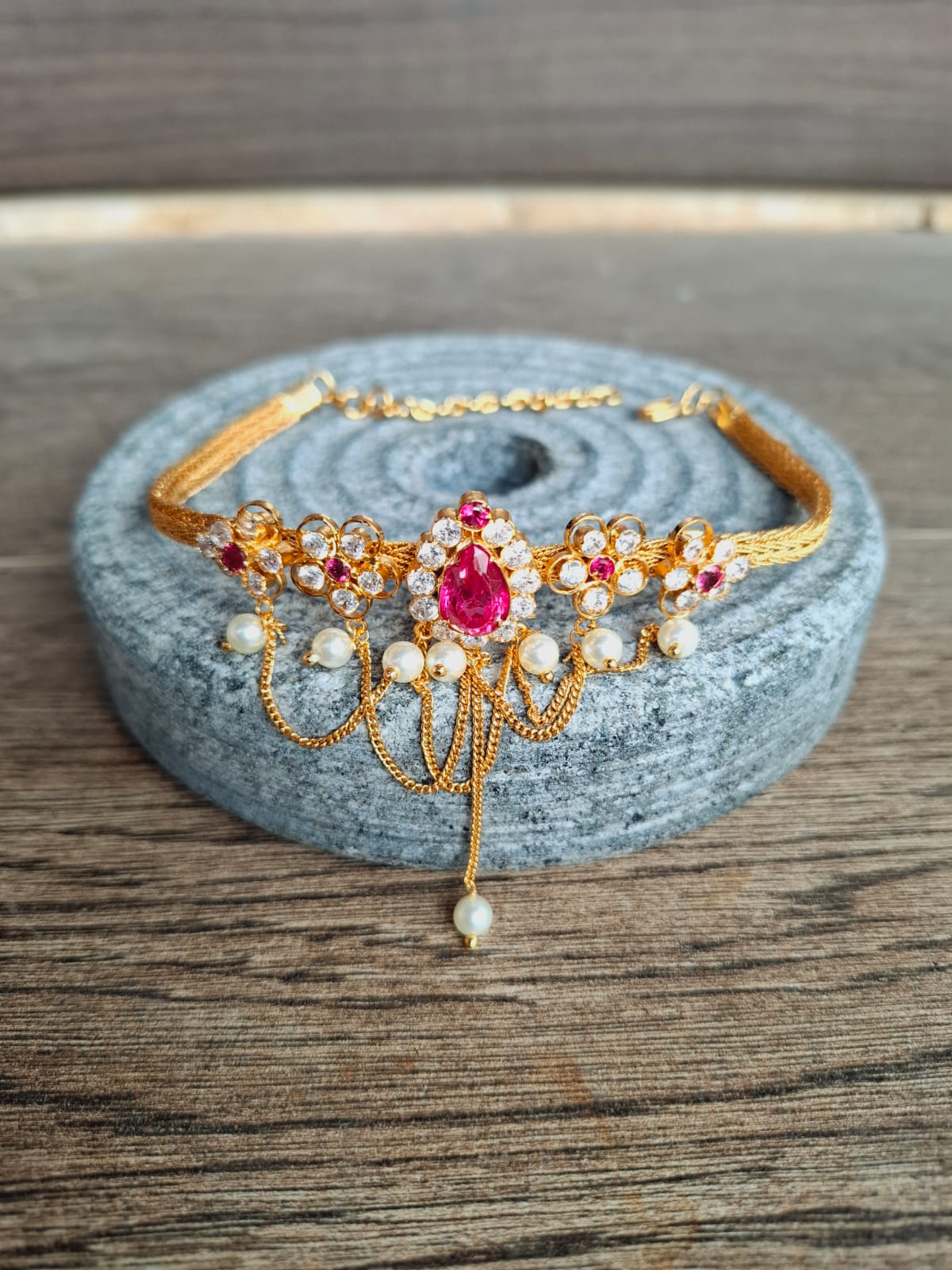 Ornate gold-toned floral choker with vibrant ruby-colored gemstones and pearls, placed on a gray stone backdrop.
