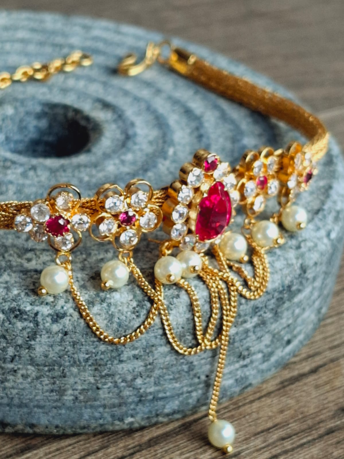 Ornate gold floral pendant choker with sparkling ruby-colored gems and delicate pearls on a textured stone backdrop.