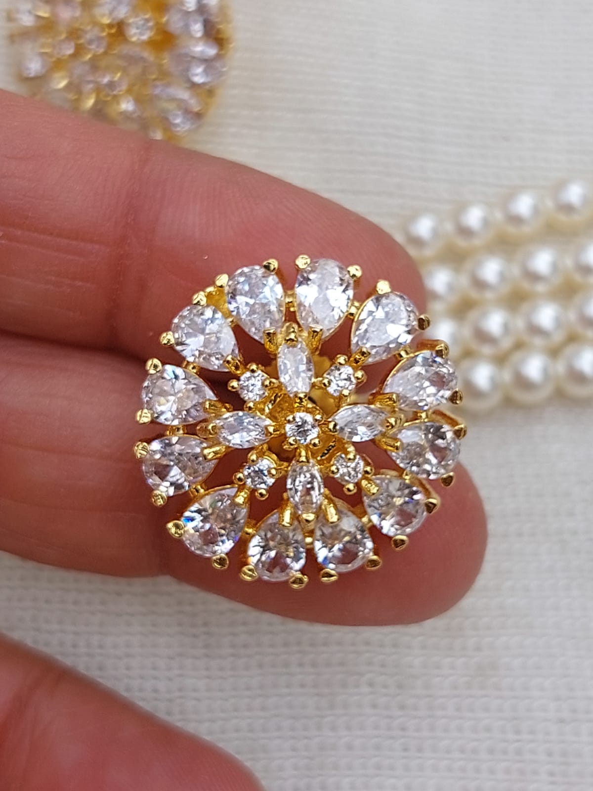 Exquisite gold-tone brooch with sparkling white stones arranged in a radiant floral design, showcased against a pearl necklace in the Mugdha Jewellery Store product image.