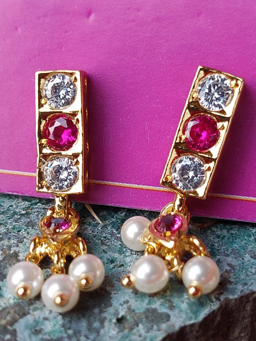 Elegant gold-tone earrings with sparkling diamonds, ruby accents, and delicate pearl clusters, displayed against a vibrant pink background.