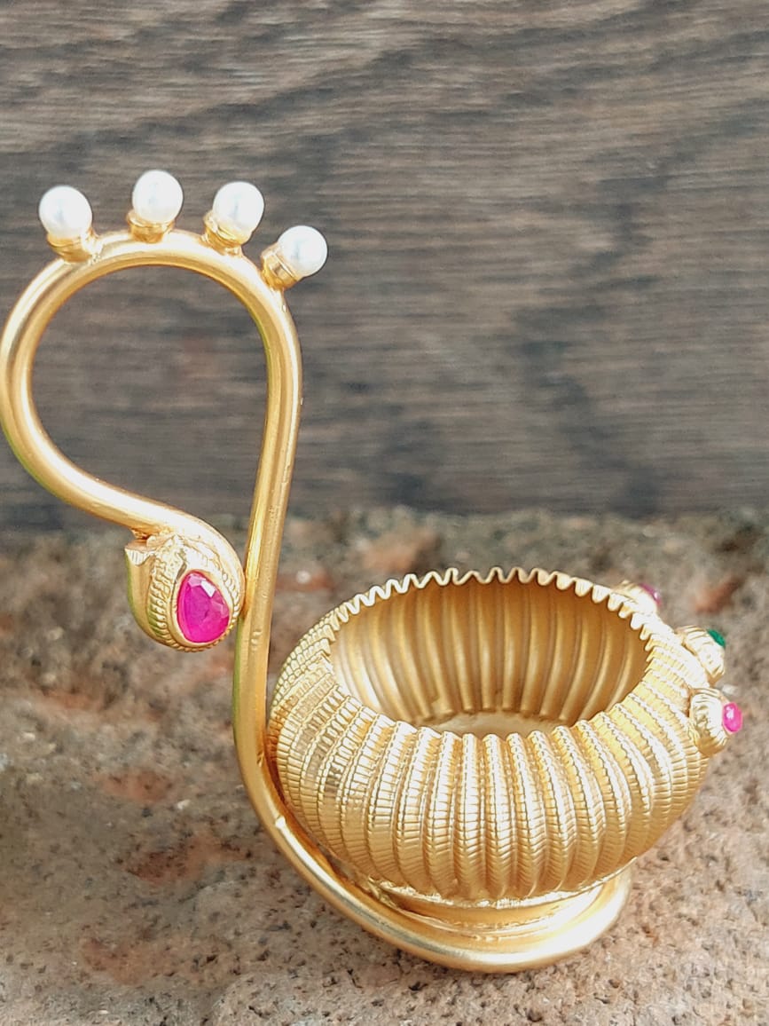 Decorative gold-toned haldi kumkum karanda holder with floral motifs and a small pink stone accent, placed on a wooden surface.