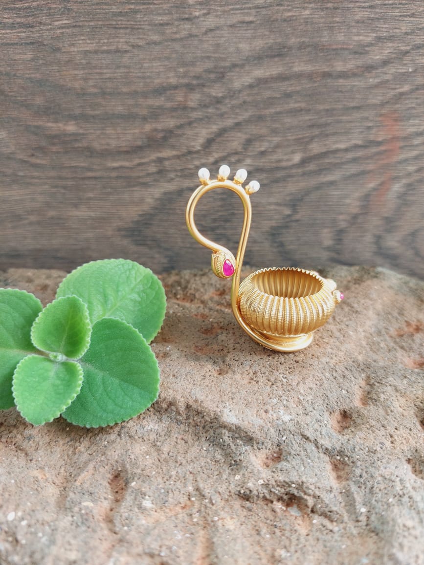 Gold-toned decorative haldi kumkum karanda trinket holder with faux pearls, displayed on a rustic wooden surface along with green plant leaves.