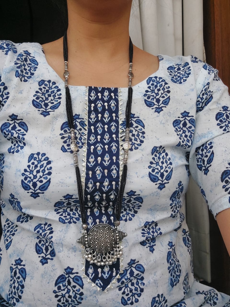 Elegant designer oxidised long mangalsutra necklace with intricate floral pattern displayed on white and blue patterned blouse.