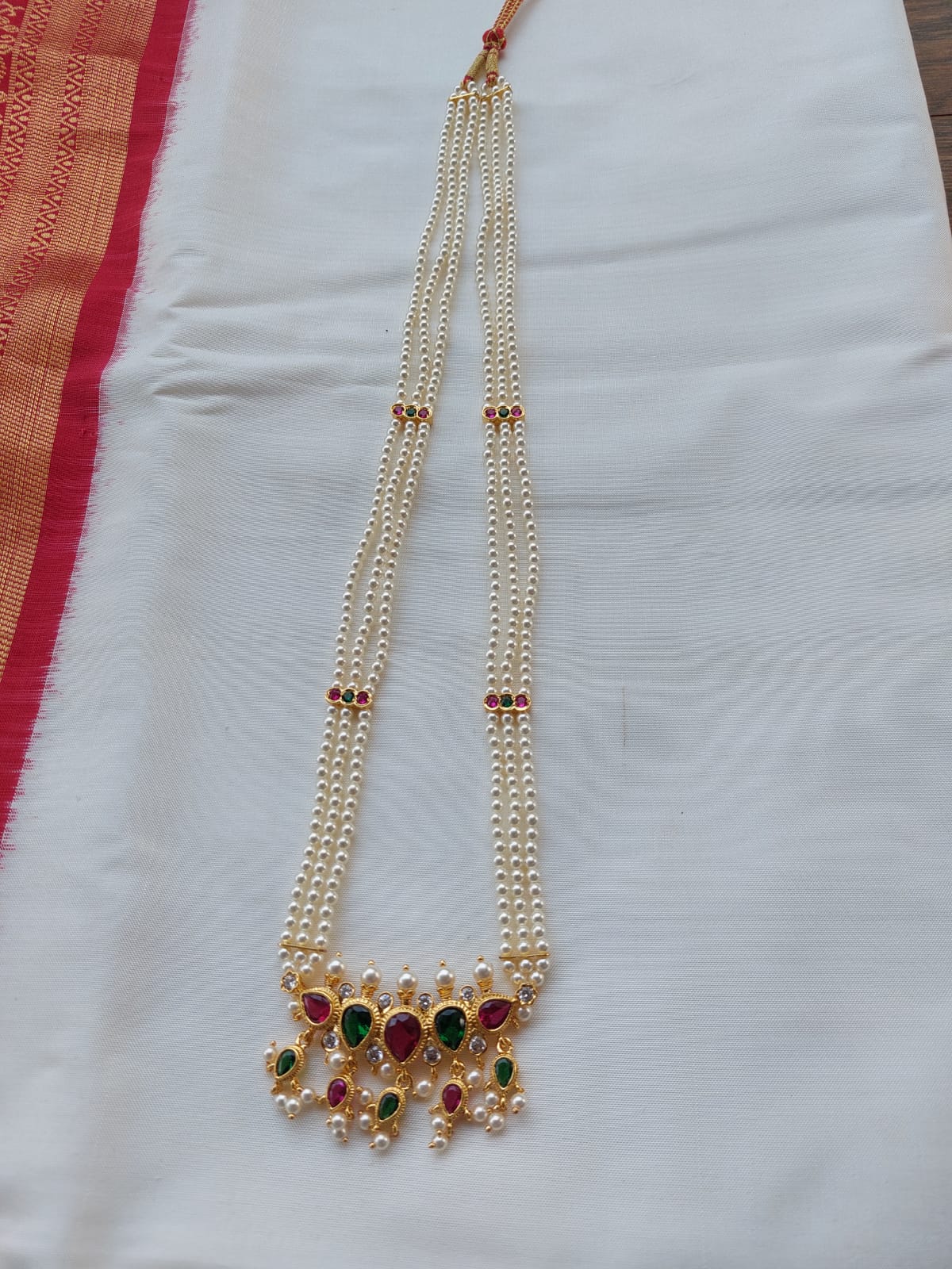 Multicoloured Long Tanmani 3 Line Haar, elegant gold-toned necklace with gemstone accents, displayed on white fabric background.