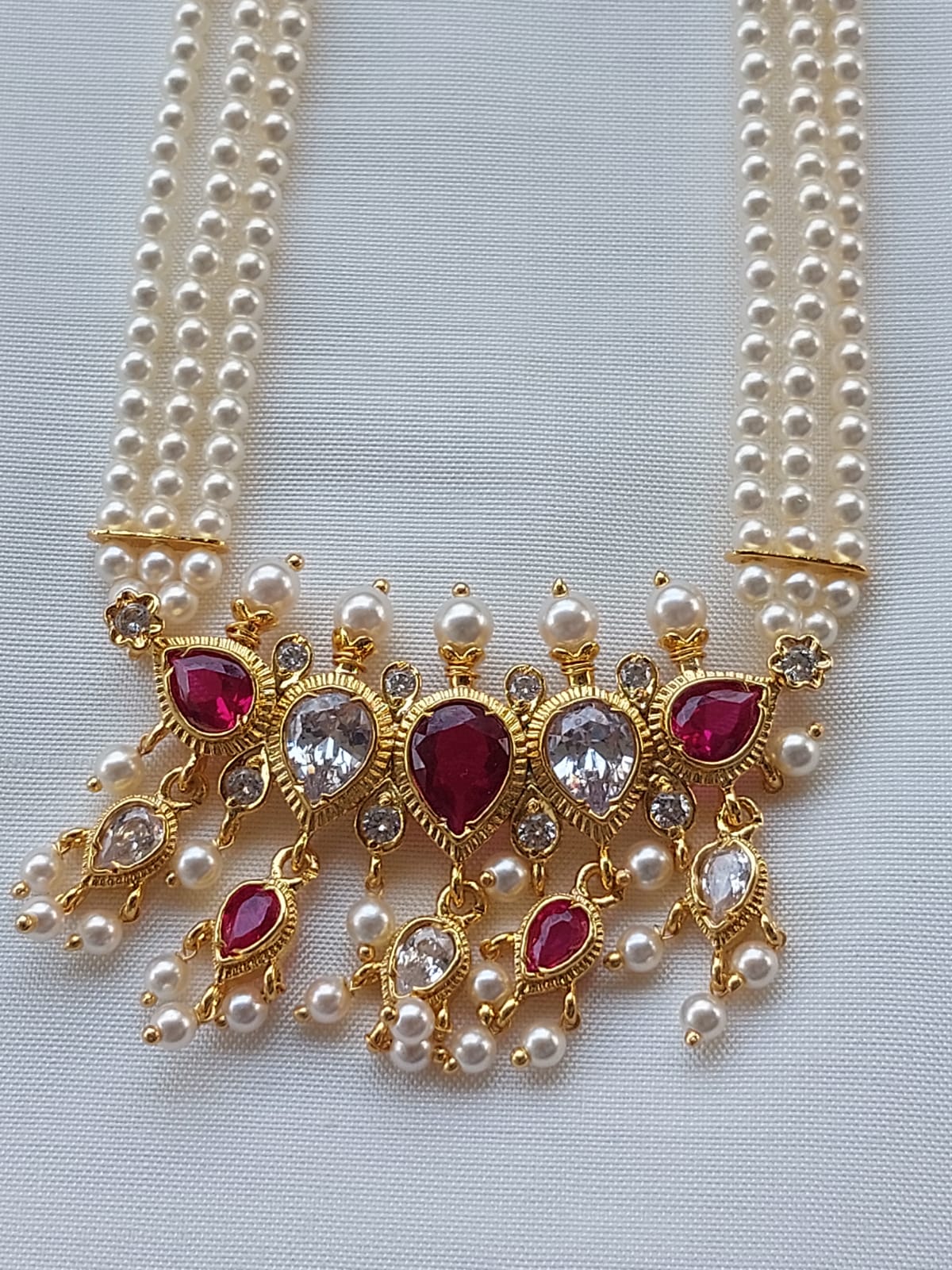 Elegant multi-line pearl necklace with ornate gold-tone pendant featuring ruby and crystal accents, showcased on a light background.