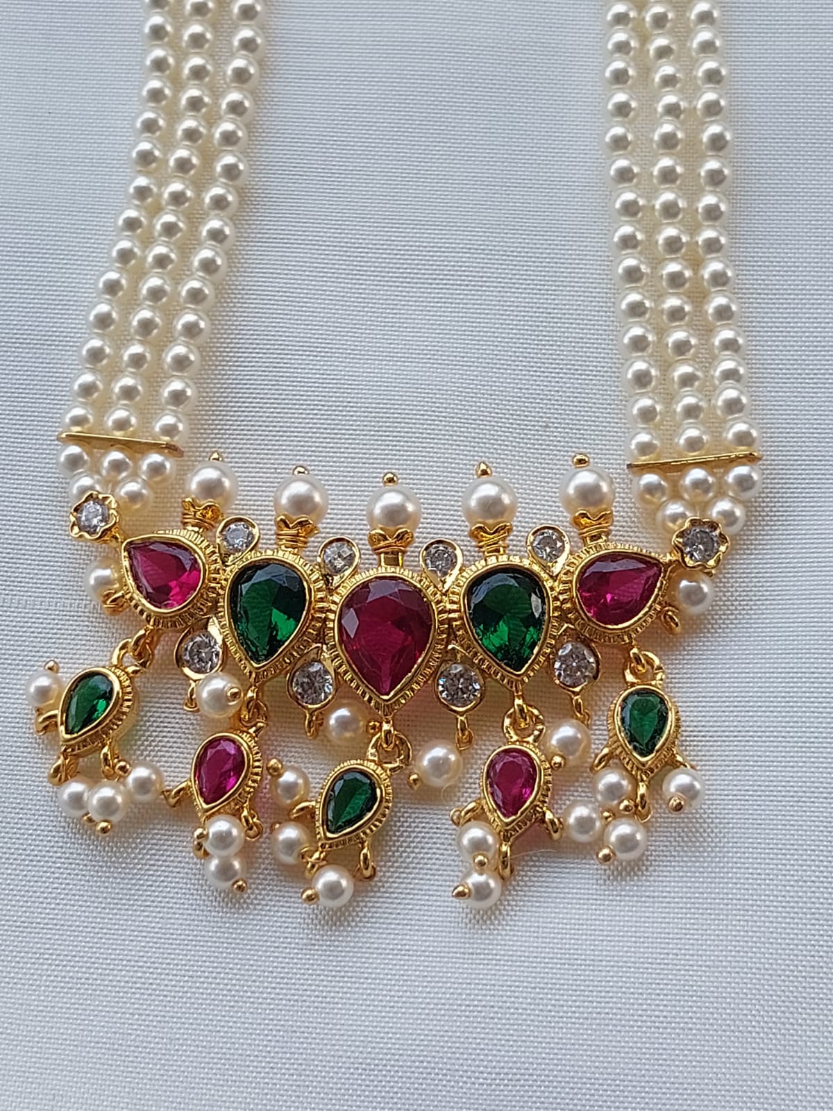 Elegant multicolored gem-encrusted pearl necklace from Mugdha Jewellery Store, featuring vibrant ruby and emerald gemstones set in a golden frame with three strands of lustrous pearls.