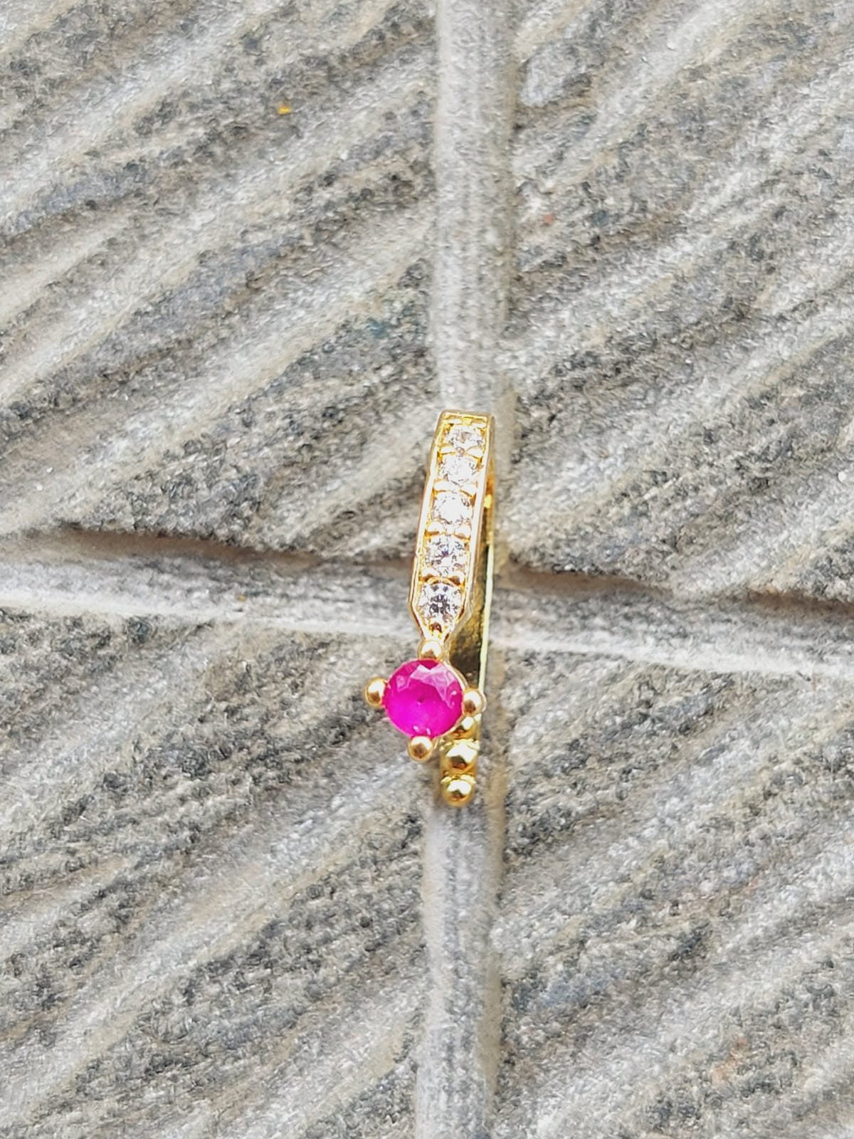Elegant gold-toned nose pin with sparkling pink gemstone center and decorative accents, placed on a textured gray background.