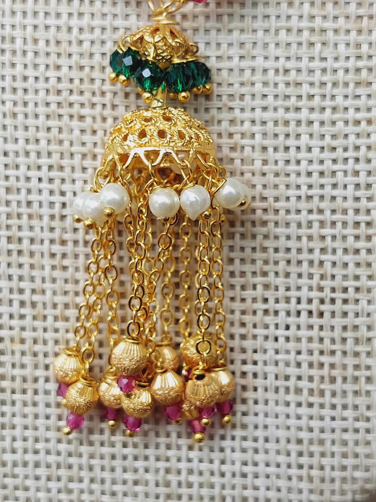 Elegant gold and emerald green designer jhumka earrings displayed on a textured fabric background from the Mugdha Jewellery Store.