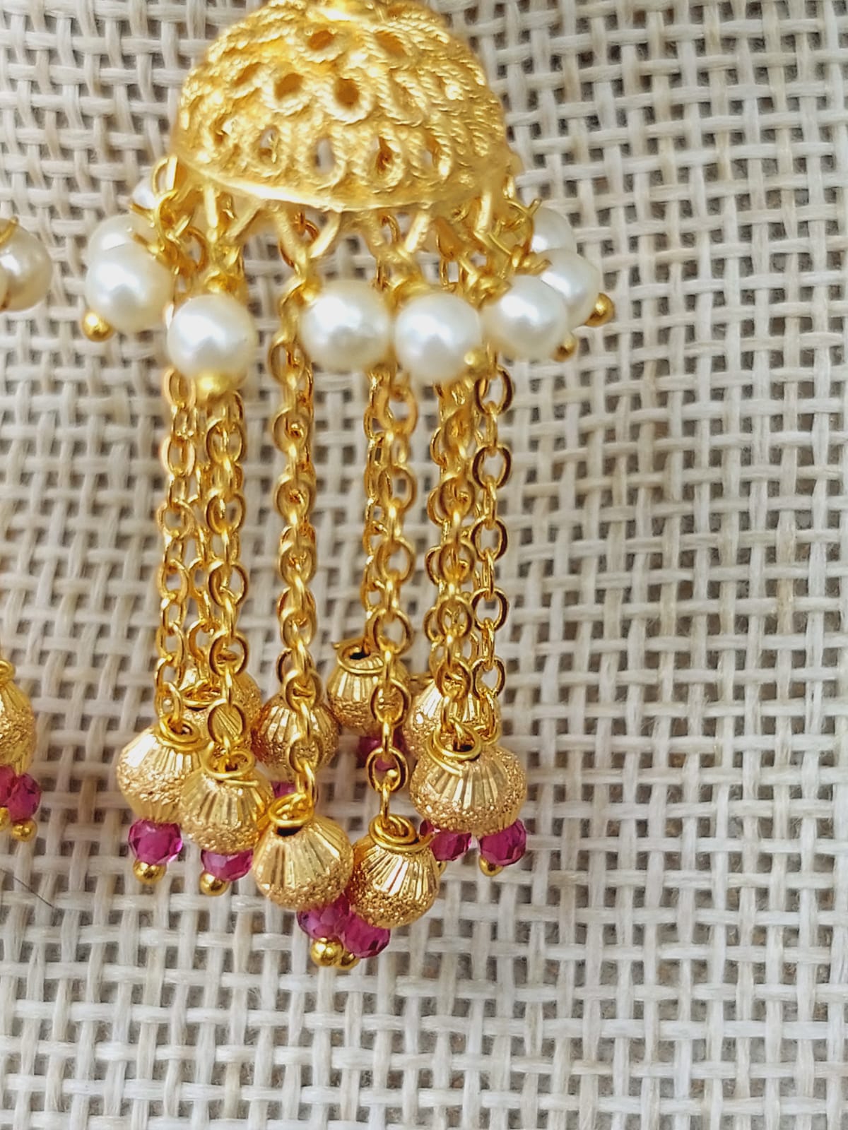Intricate golden earrings with a floral design, accented with delicate pearls and ruby-colored embellishments, showcased against a light-colored textured background.