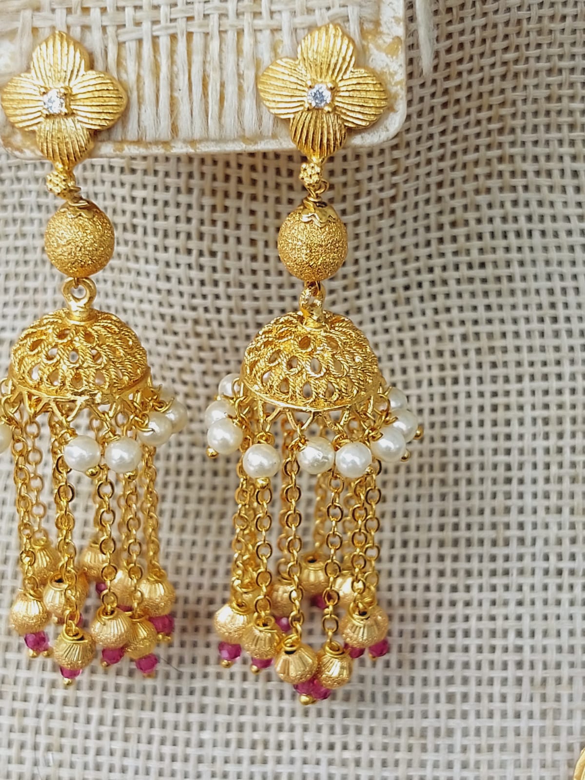 Elegant gold-toned Nandini Zhumki designer earrings with intricate floral patterns, dangling pearl and bead accents, showcased against a woven backdrop.