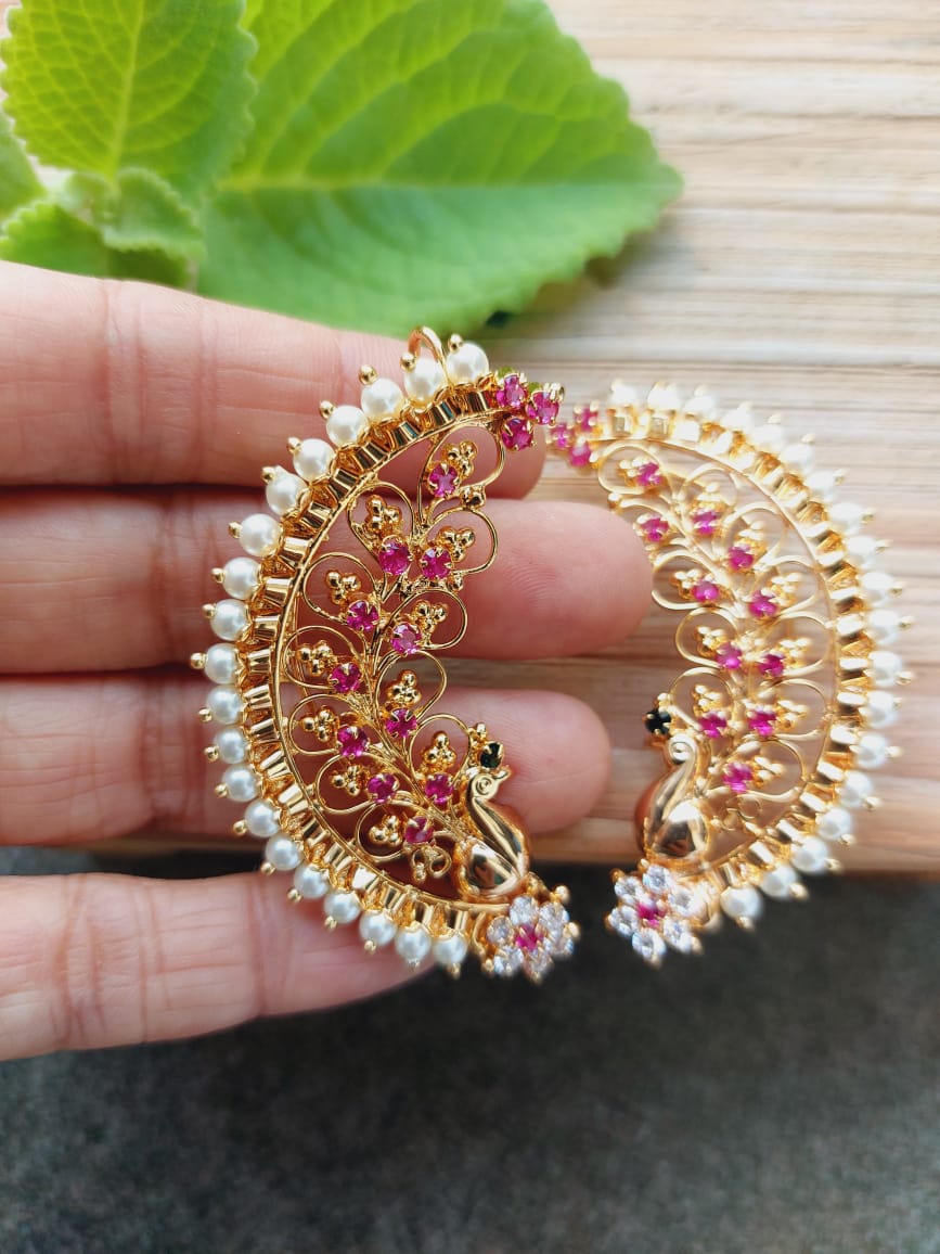 Ornate gold-toned earrings with intricate floral and pearl designs, held in a hand against a green leaf backdrop.