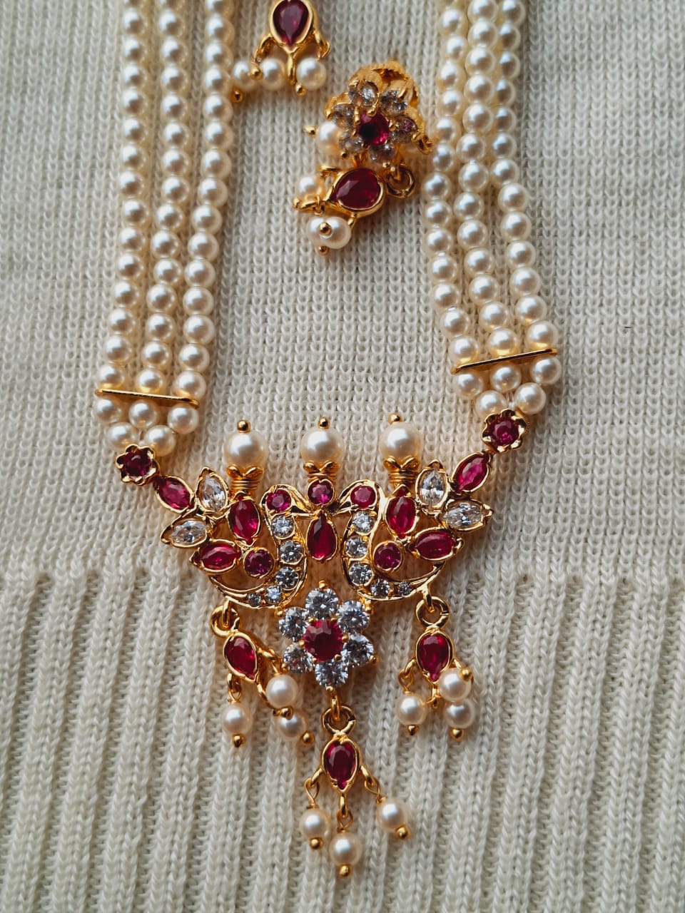 Ornate Peacock Pendant with Pearls and Jewels
This image shows an intricate peacock-shaped pendant made of gold-toned metal, featuring a cluster of sparkling red and clear jewels and adorned with multiple strands of lustrous pearls. The pendant is the focal point of the piece, surrounded by the delicate pearl necklace.