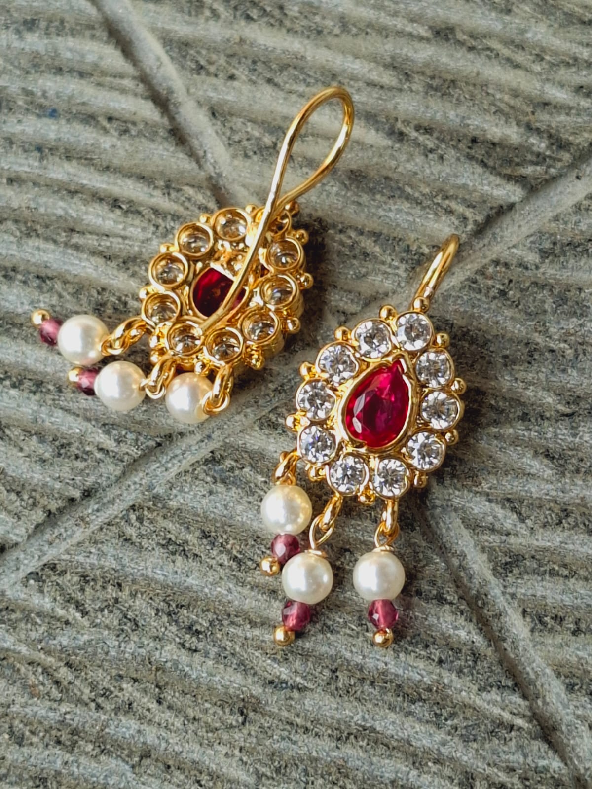 Elegant gold-tone designer earrings with red teardrop gems, sparkling rhinestones, and pearl accents, displayed on a textured gray background.