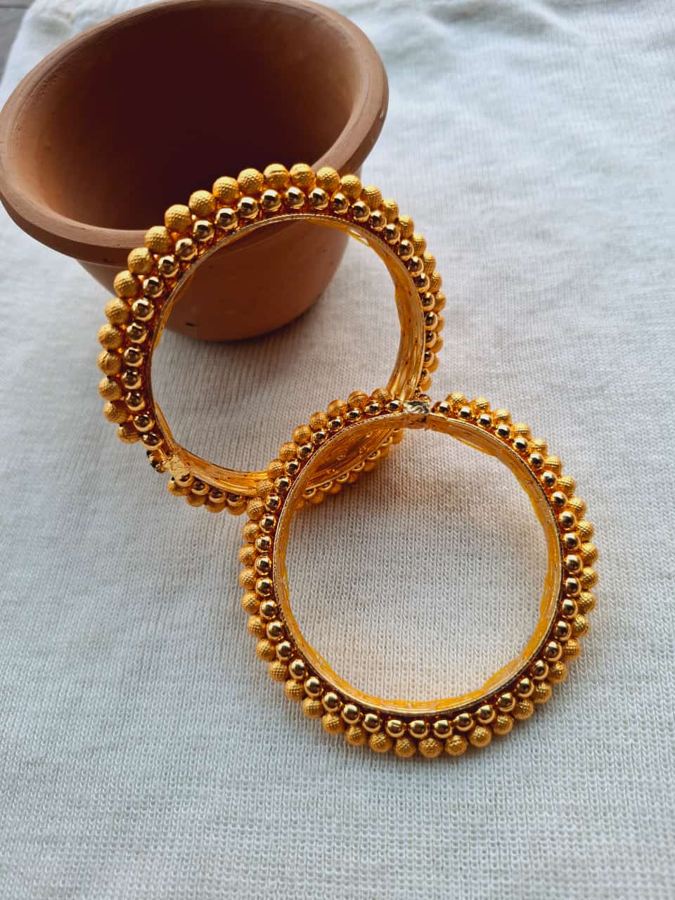 Ornate golden bangles in a traditional Indian style, featuring intricate beaded detailing and a decorative ceramic container in the background, showcasing the rich cultural heritage and craftsmanship of Mugdha Jewellery Store.