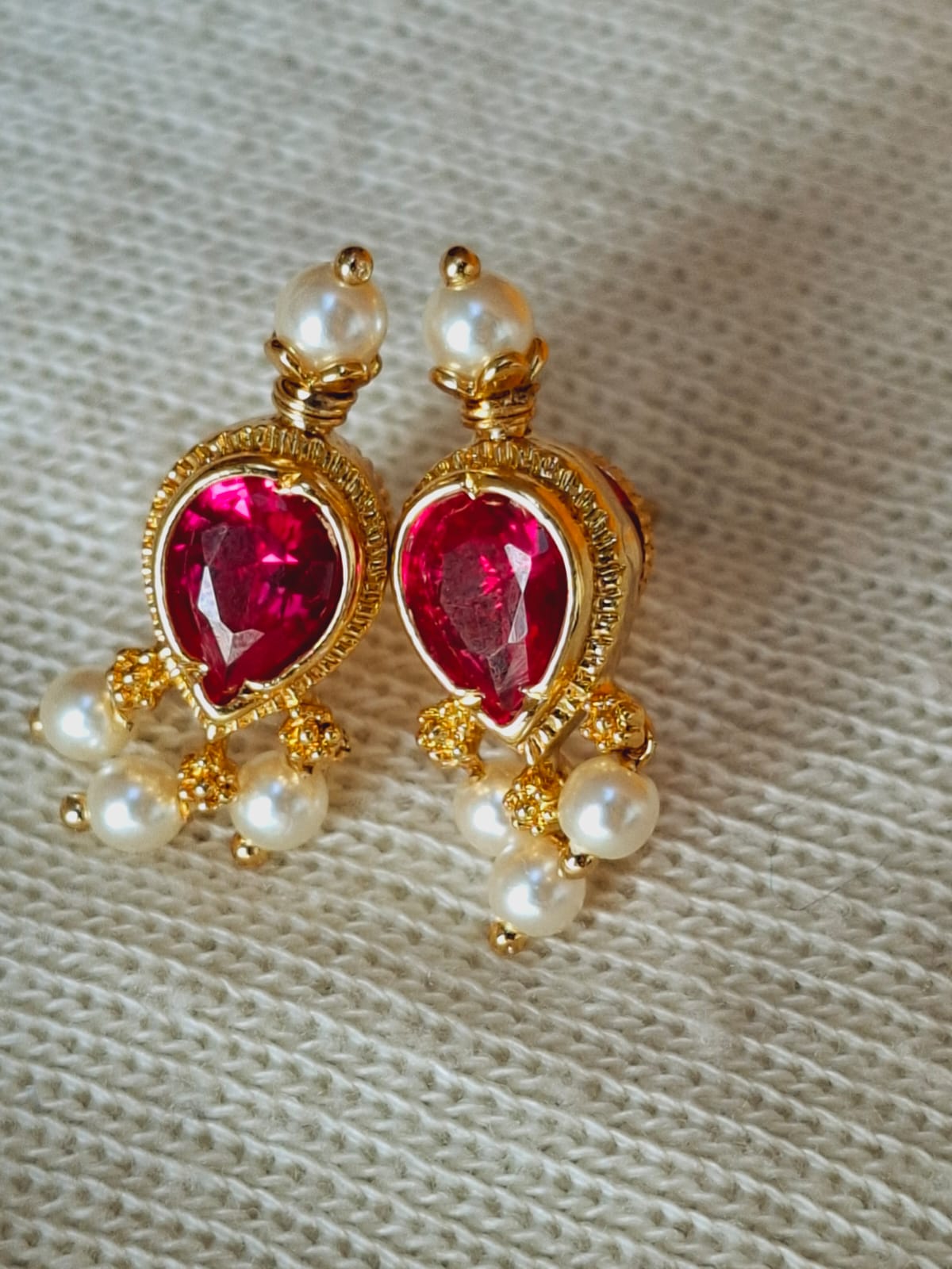 Elegant gold-tone Tanmani earrings with vibrant red gemstones and lustrous pearls, displayed on a soft gray fabric background.