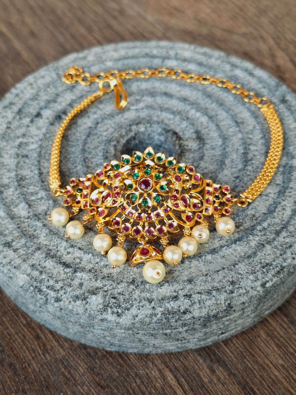 Elegant gold-toned designer bajuband adorned with vibrant gemstones and lustrous pearls, showcased on a textured gray surface.