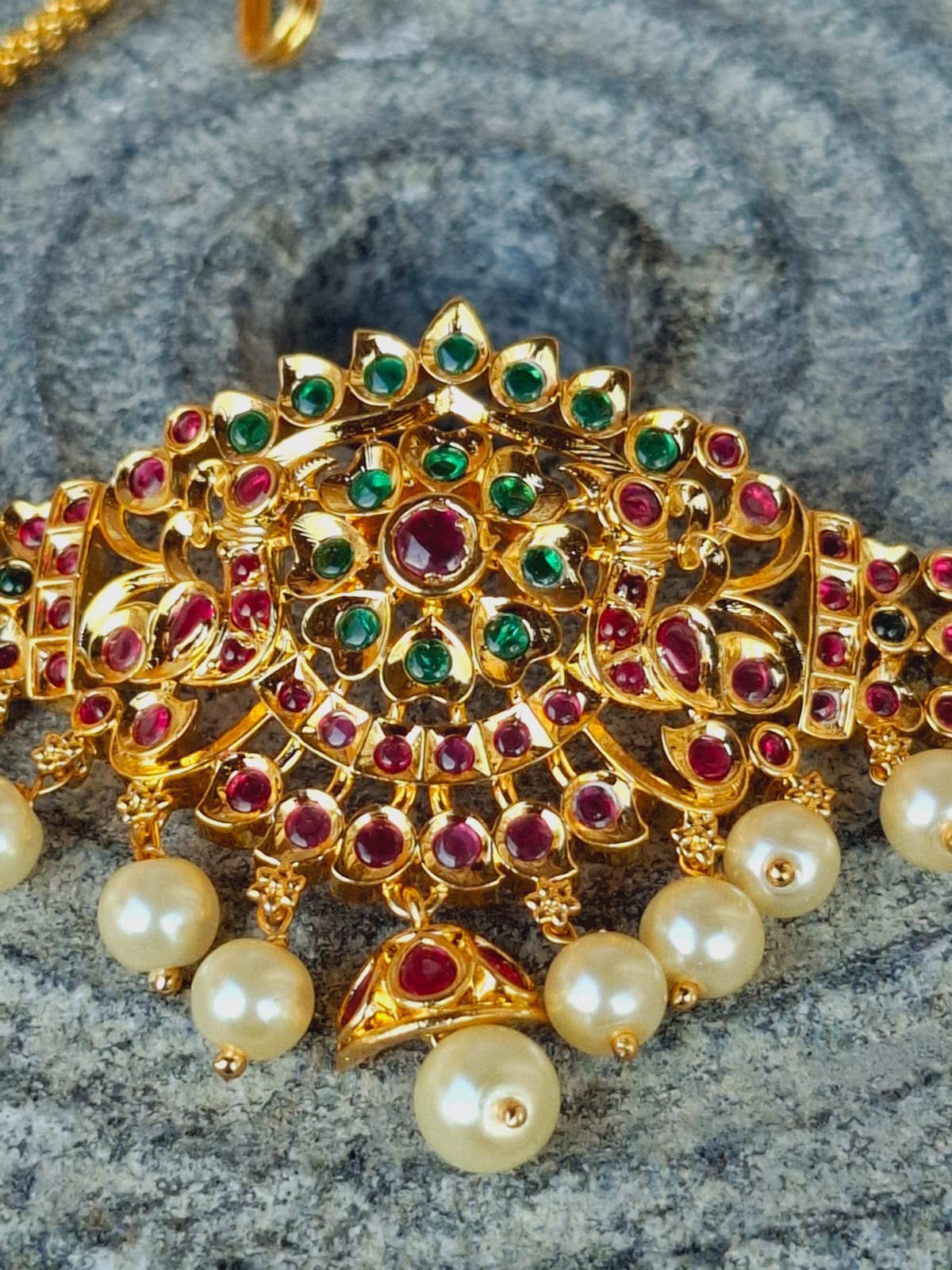 Ornate golden designer bajuband with intricate floral motif adorned with vibrant red and green gemstones, and pearls, showcased on a textured gray surface.