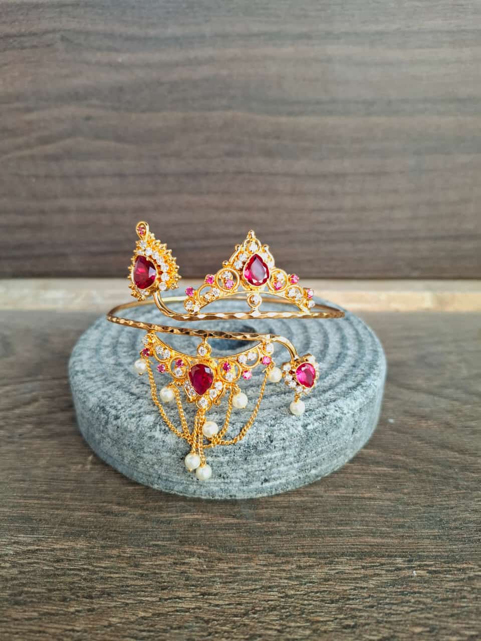 Ornate golden bajuband with intricate floral patterns and vibrant ruby-colored gems displayed on a neutral stone pedestal.