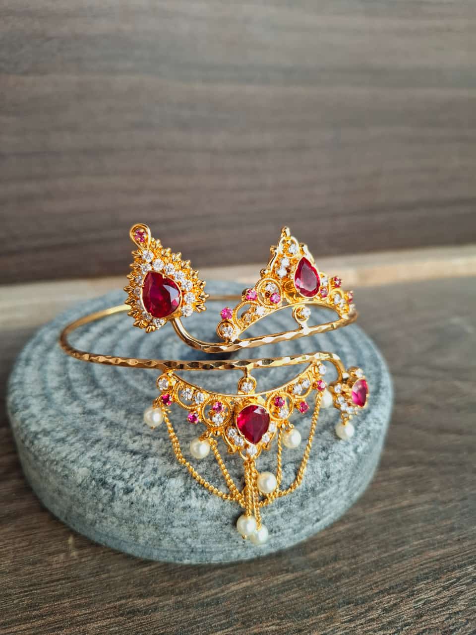 Ornate gold-toned bajuband (arm ornament) with sparkling red and orange gemstones, displayed on a gray stone surface.