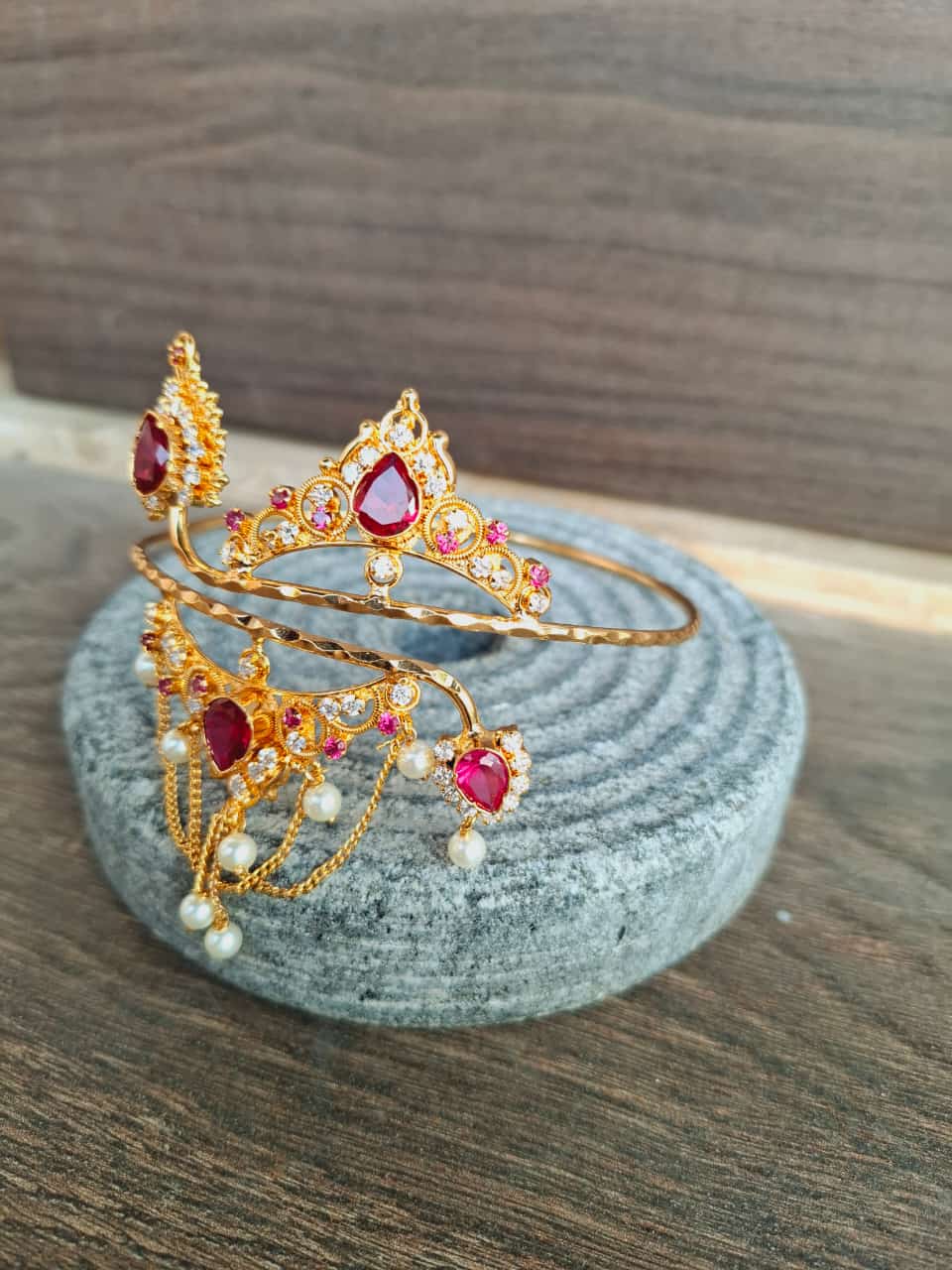 Elegant gold-toned Samradnyi Bajuband adorned with vibrant ruby-colored gemstones, displayed on a textured gray surface.