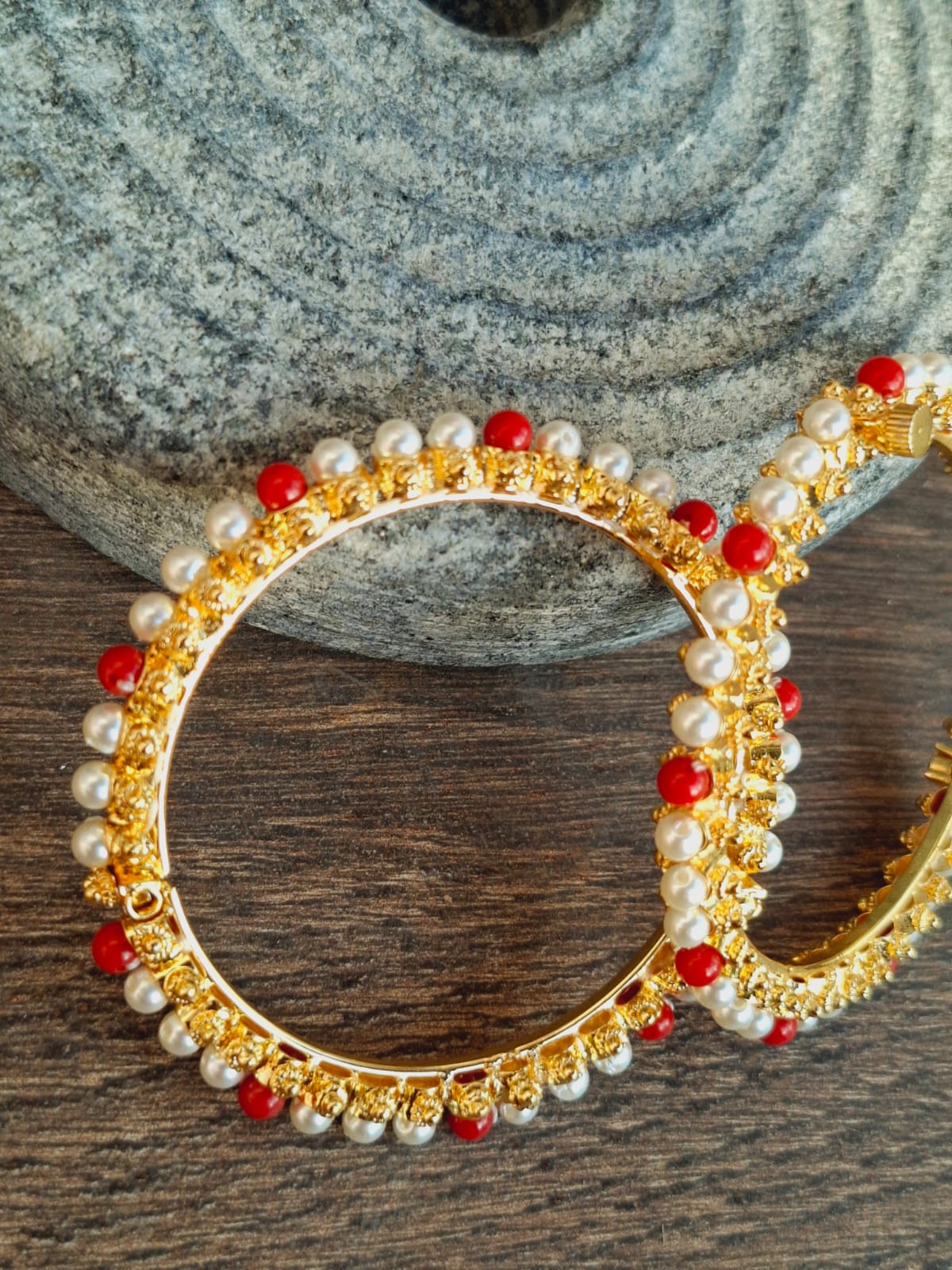Elegant golden bangle with vibrant red stones and shimmering pearls displayed on a textured gray background and wooden surface.