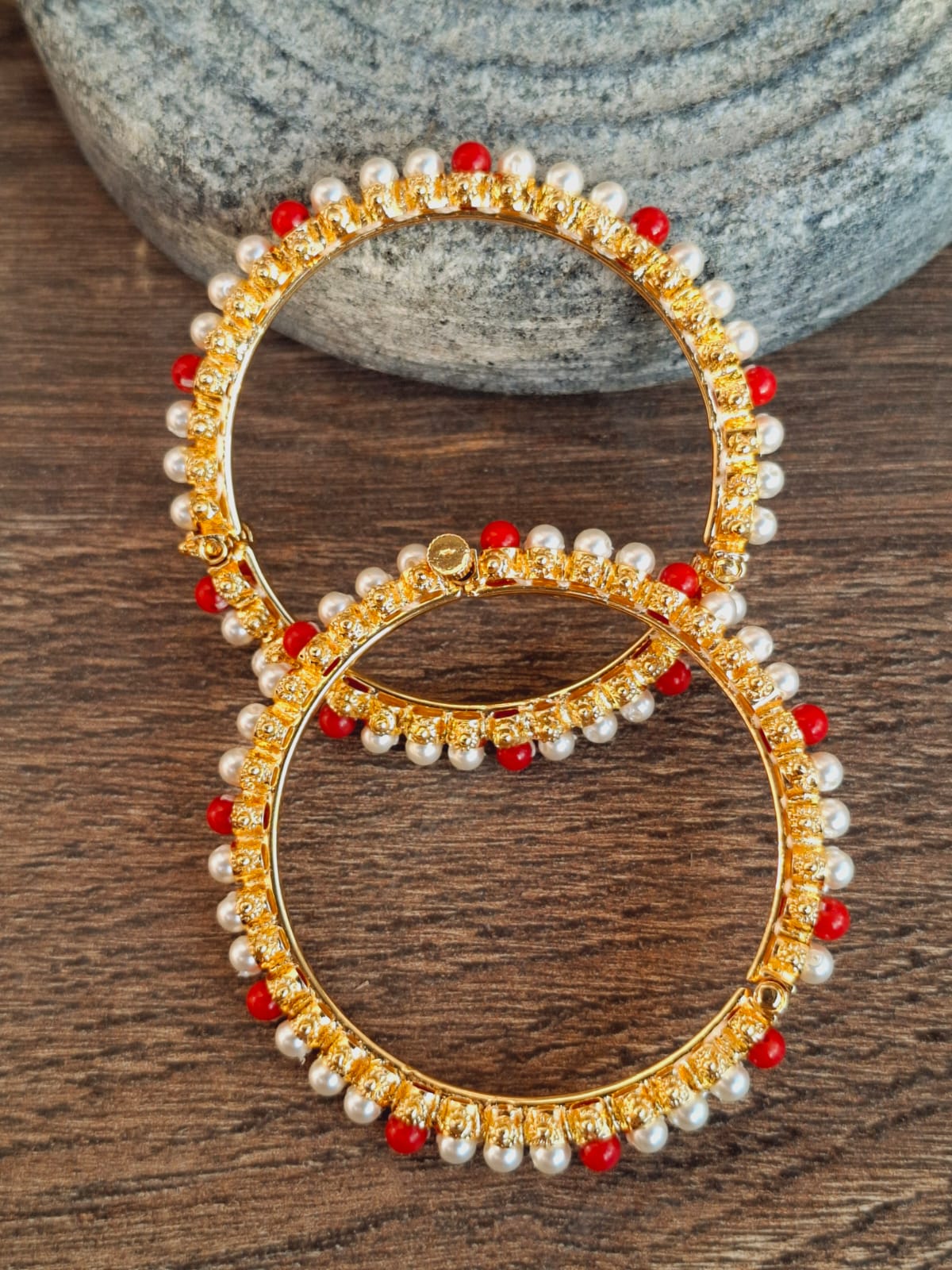Gold-tone pearl and red stone encrusted earrings on a wooden surface with a natural stone backdrop.