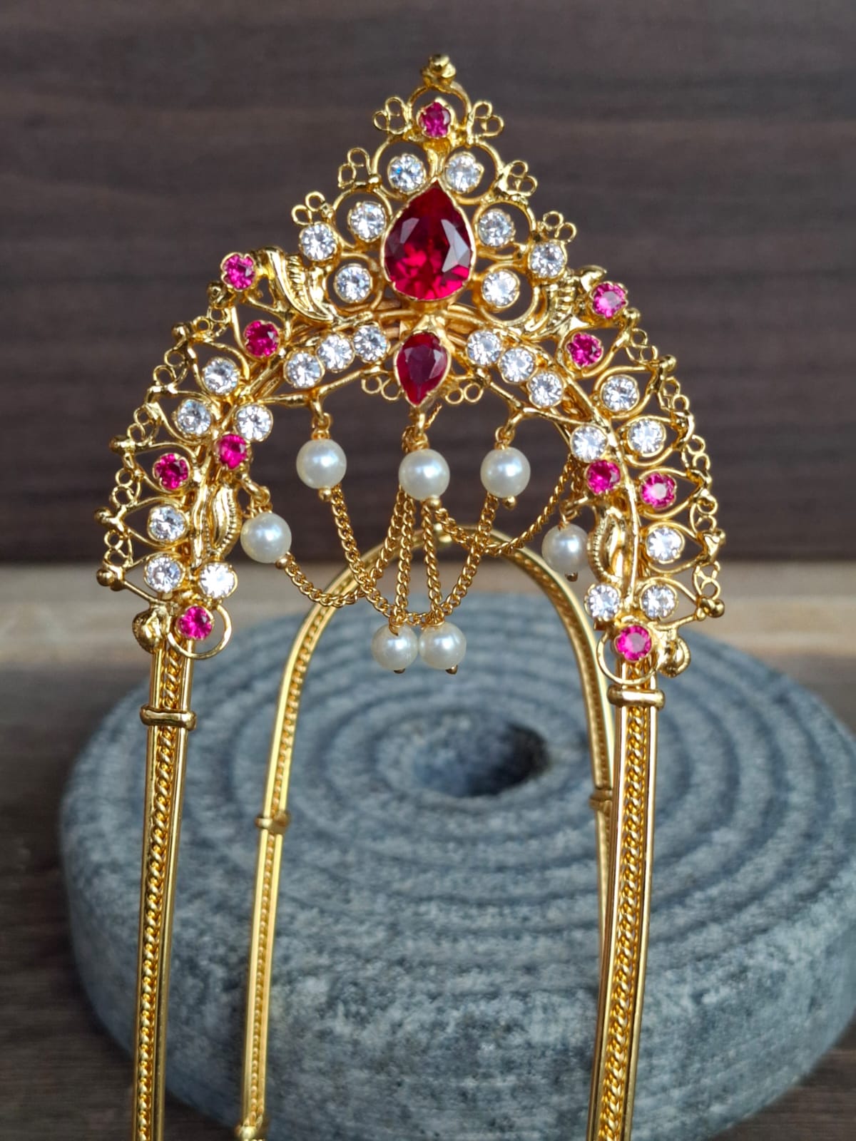 Ornate gold and gemstone jewelry piece displayed on a gray background. The piece features a crown-like design with intricate metalwork and various colored gemstones, including red and white gems. The pendant appears to have hanging pearl accents, adding to the opulent and regal appearance of the Mhalsa Bajuband from the Mugdha Jewellery Store.