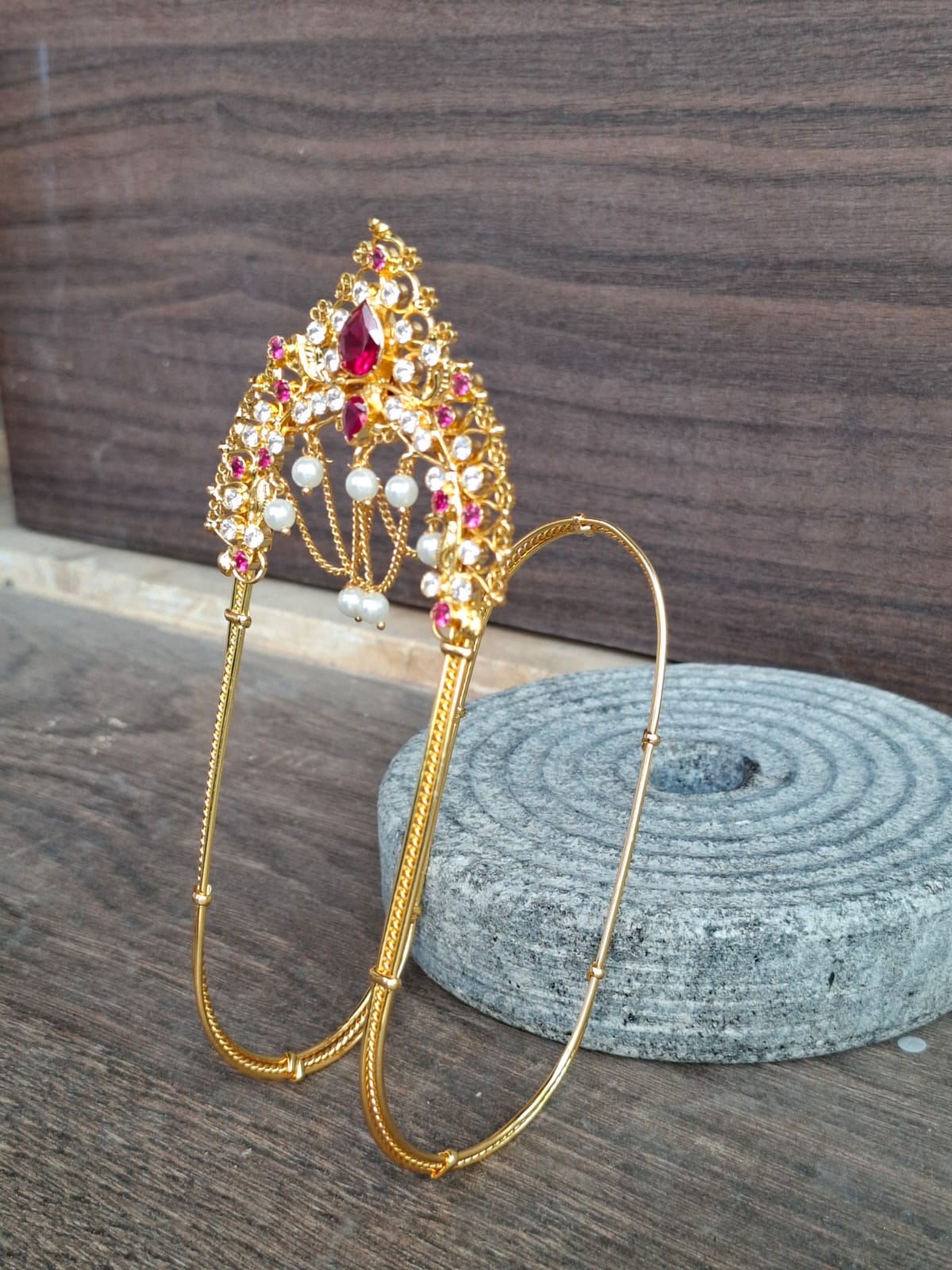 Ornate gold-toned Mhalsa Bajuband with vibrant red and colorful gemstones, showcased on a wooden backdrop.