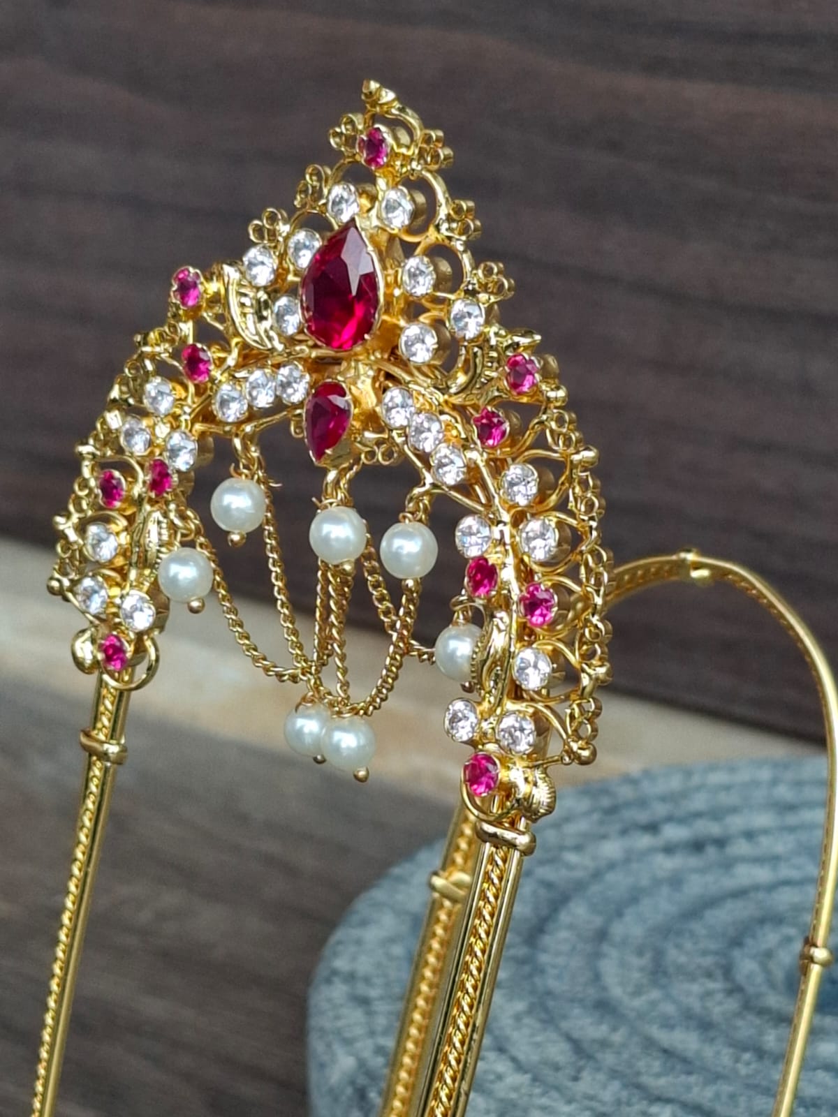 Ornate gold-toned Mhalsa Bajuband with sparkling red and white gems, featuring a prominent teardrop-shaped centerpiece and decorative dangling chains, displayed on a textured background.