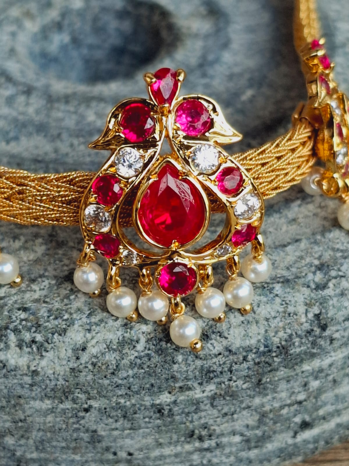 Ornate gold and ruby-colored peacock choker necklace with pearl accents displayed on a grey marble surface