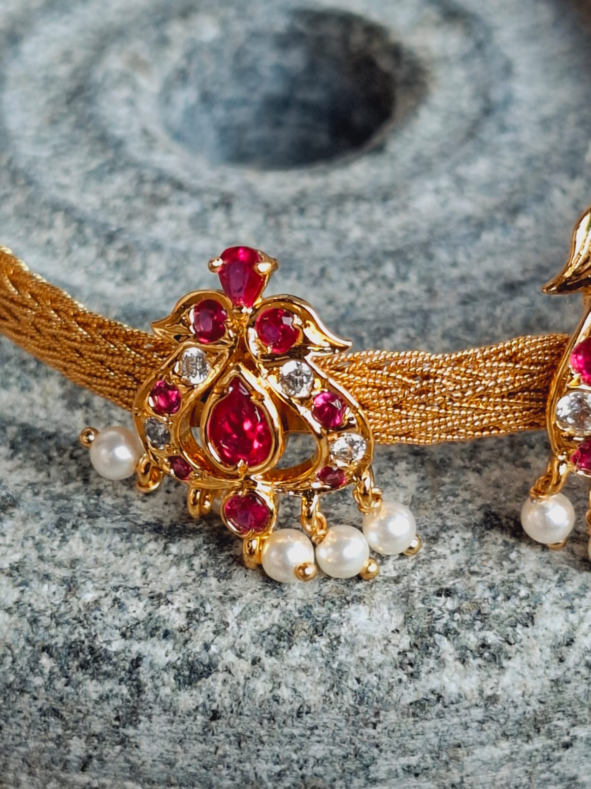 Intricate gold-toned Chatai Peacock Chocker cum Bajuband with vibrant red gemstones and delicate pearls against a grey stone backdrop
