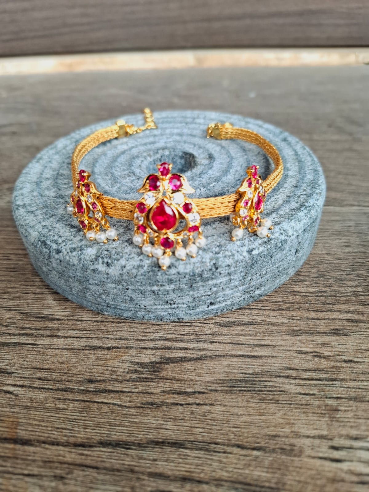 Chatai Peacock Chocker cum Bajuband, a stunning gold-toned jewelry set featuring intricate peacock designs and vibrant red and white gemstones, displayed on a gray textured surface.