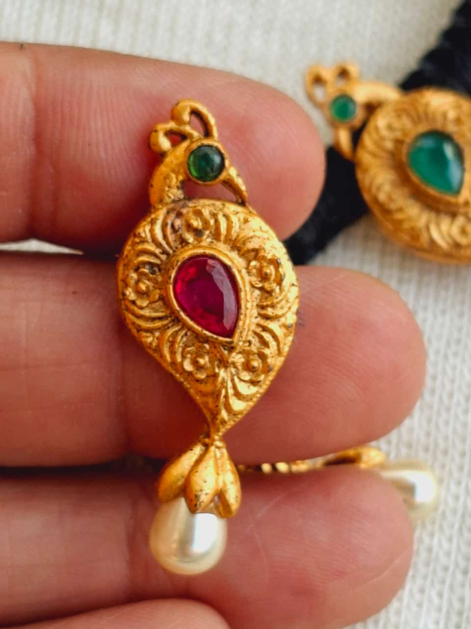 Ornate golden mangalsutra pendant with intricate floral patterns, bezel-set ruby stone, and hanging pearl accent, showcased against a neutral background at the Mugdha Jewellery Store.