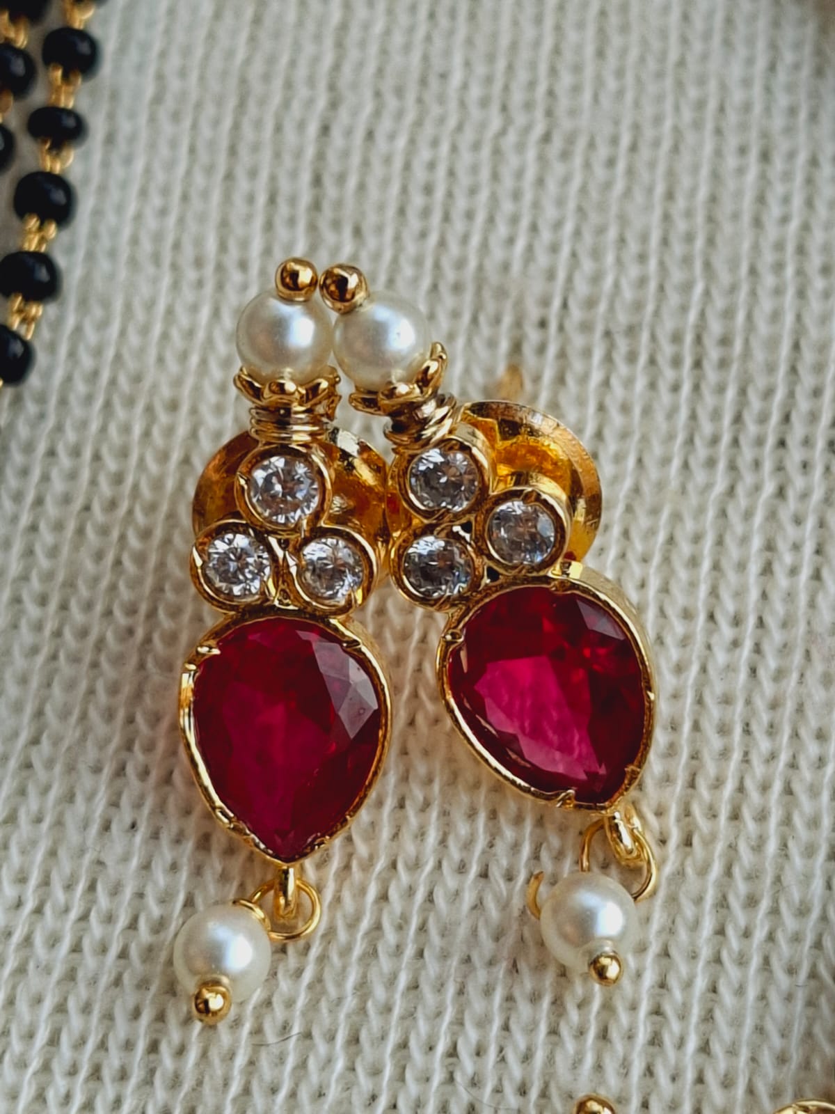 Elegant multicolored mangalsutra with gold-toned metal, ruby-hued gemstones, and lustrous pearls on a knitted textile background.