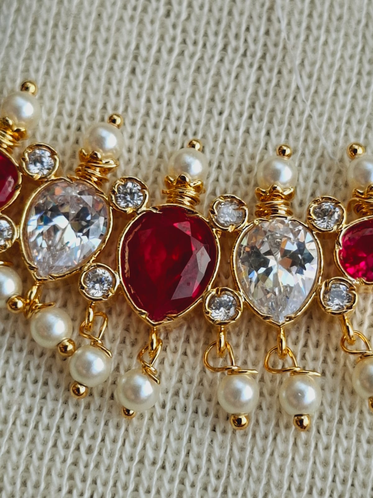 Ornate gold-toned mangalsutra with red, white, and diamond-like stones, featuring dangling pearls, showcased on a textured off-white fabric background.
