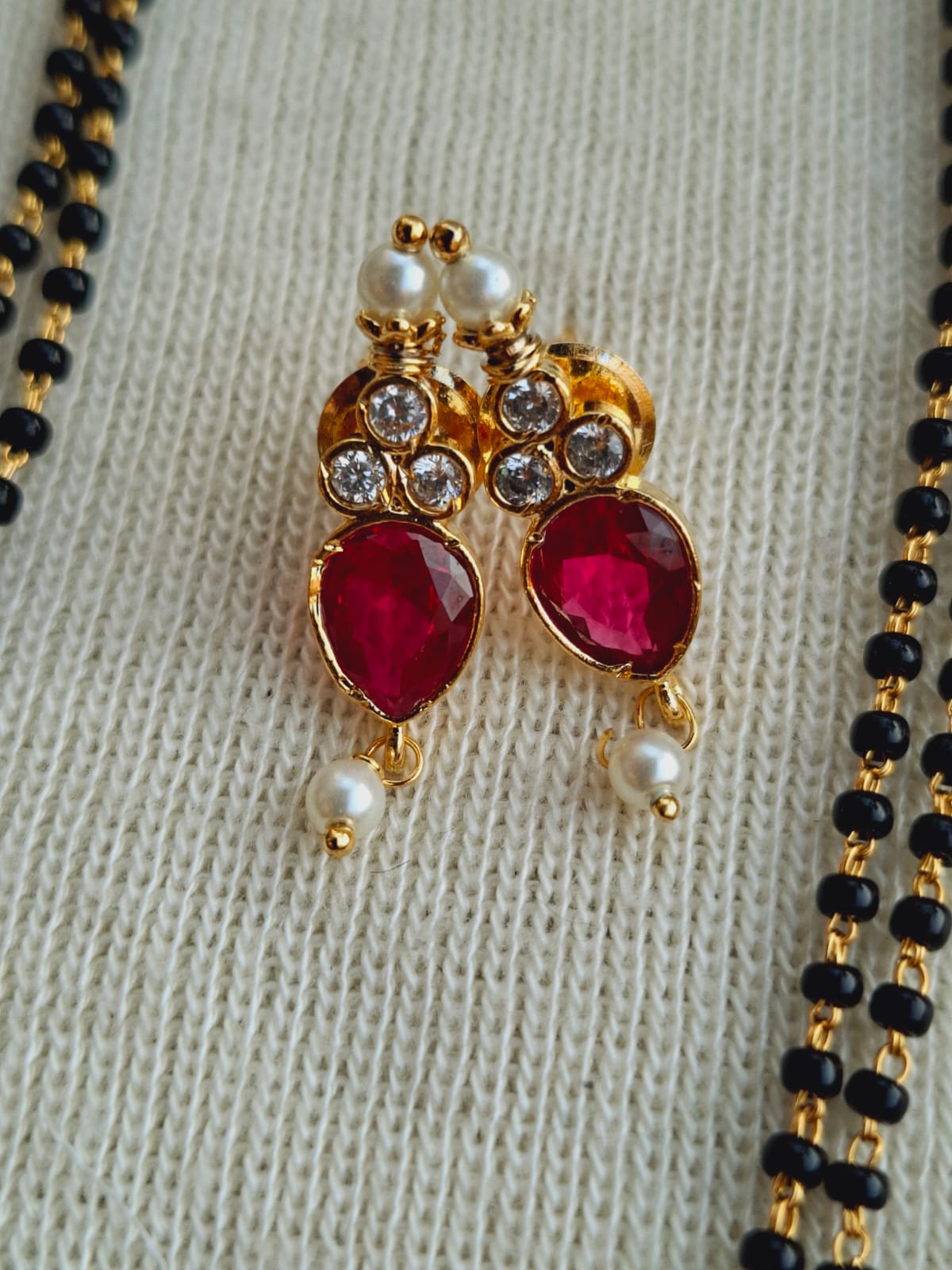 Elegant gold-toned mangalsutra pendant with red gemstones and white pearls, showcased against a neutral fabric background.