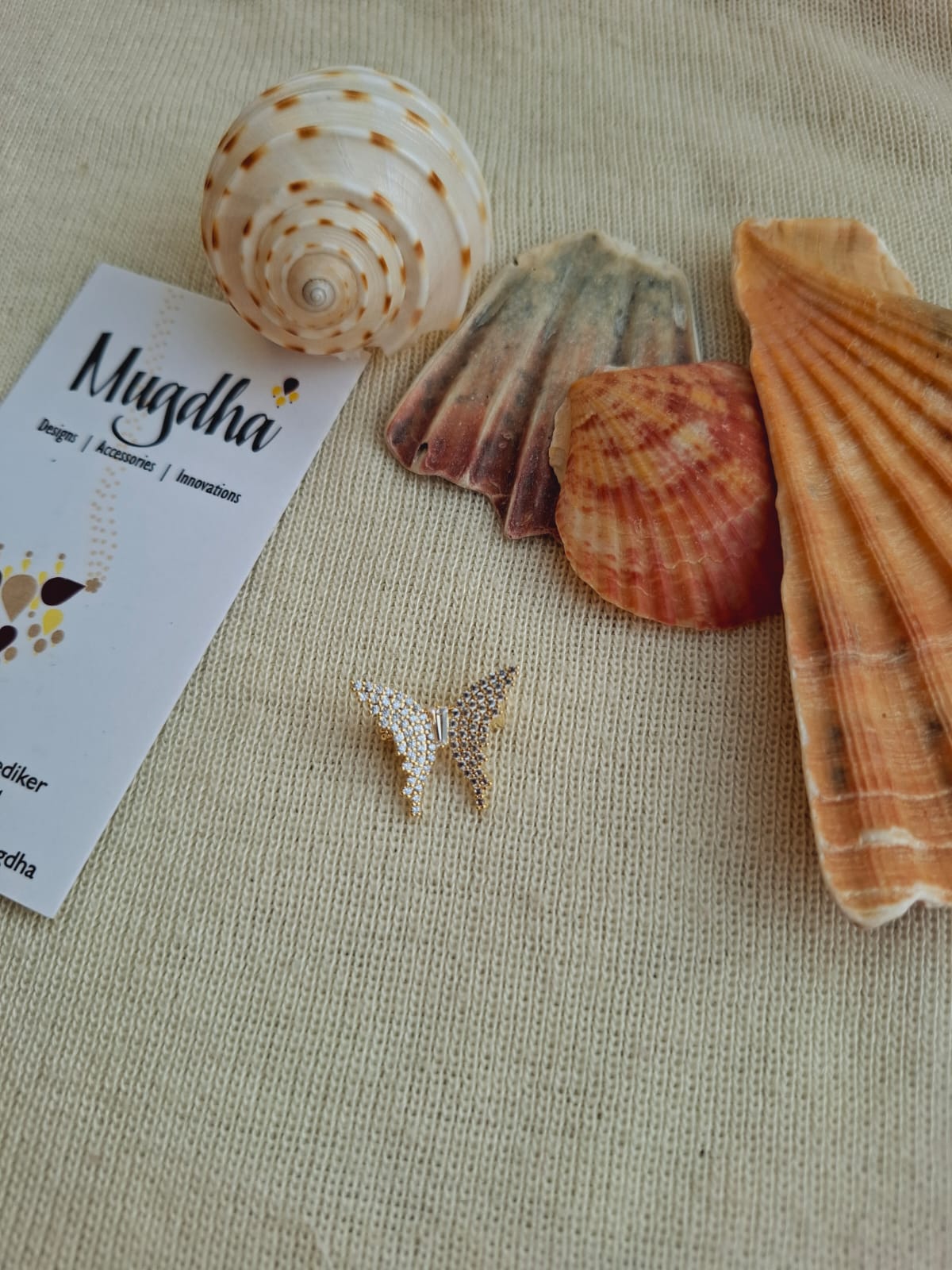 Beautiful butterfly-shaped saree pin brooch from Mugdha Jewellery Store, displayed with seashells and fabrics on a tan background.