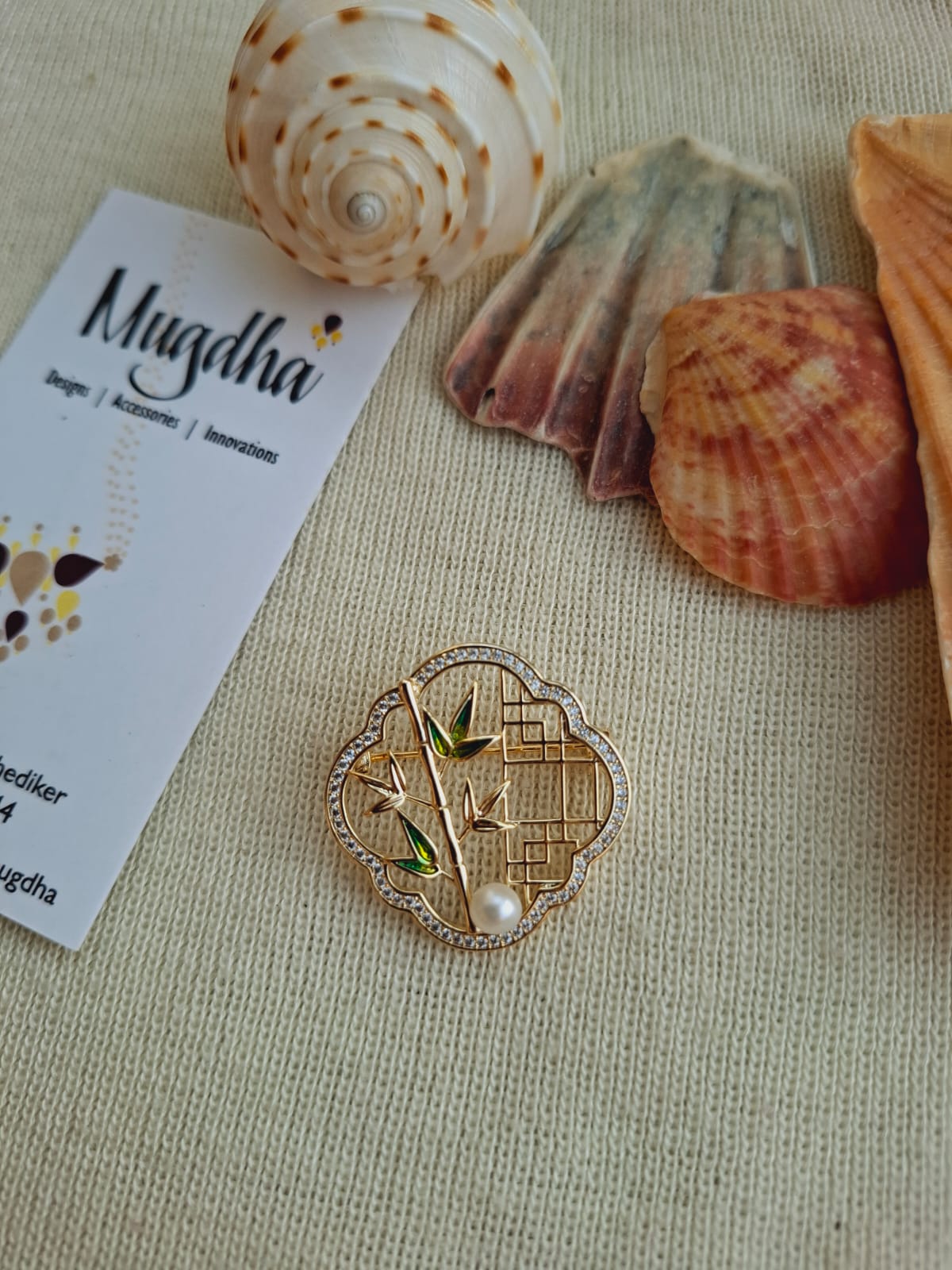 Elegant bamboo design saree pin and brooch by Mugdha Jewellery Store, surrounded by seashells and accessories on a neutral background.