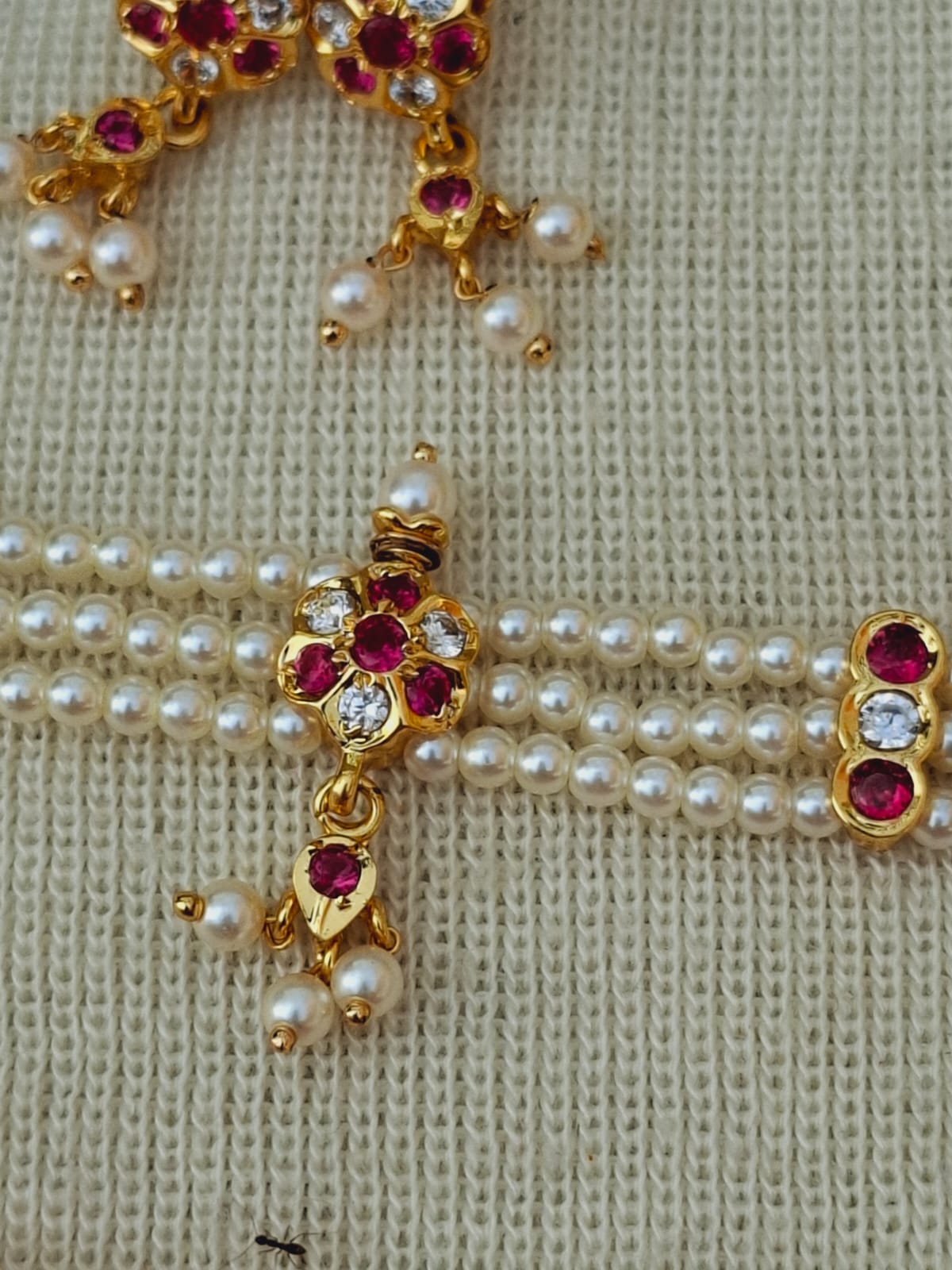 Elegant gold-toned cross pendant adorned with red rubies, white pearls, and sparkling rhinestones on a woven textured background from Mugdha Jewellery Store.