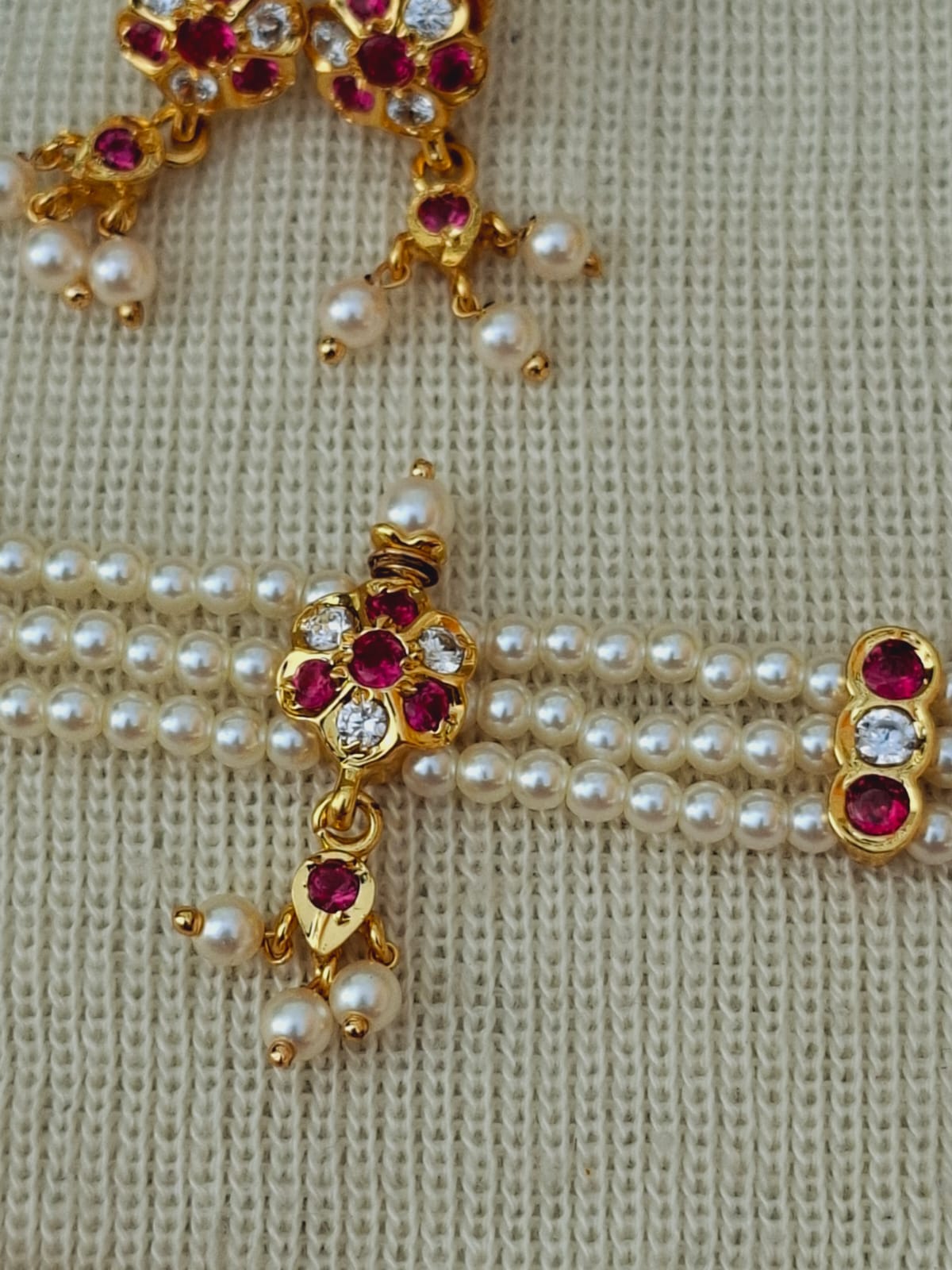Elegant gold-tone flower pendant adorned with sparkling red gemstones and lustrous pearls on a delicate pearl necklace, showcased on a textured white background.